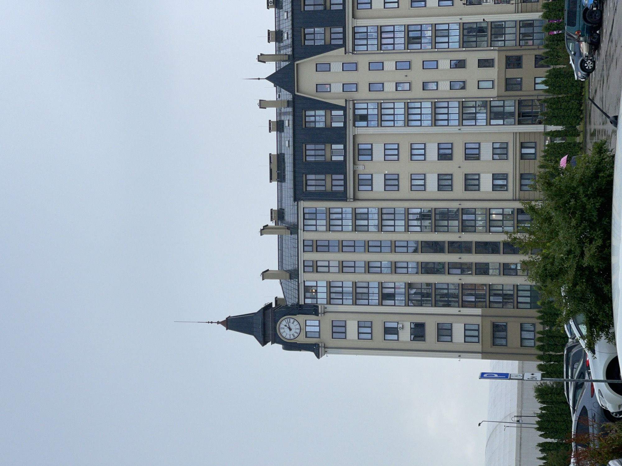 In Zelenogradsk, Russia, a modern, multi-story residential building with a distinctive clock tower dominates the frame against an overcast sky. The building features a light-colored facade with numerous windows, many of which reflect the cloudy sky. The clock tower, a prominent architectural element, has a dark, pitched roof and a visible clock face. Below the building, a parking lot is visible with several parked cars, including sedans and SUVs. A green hedge runs along the base of the building, and some trees are present, with one bush partially obscuring the lower part of the scene. A blue parking sign is visible on the left side of the image. The overall impression is of a contemporary urban environment under a muted sky.