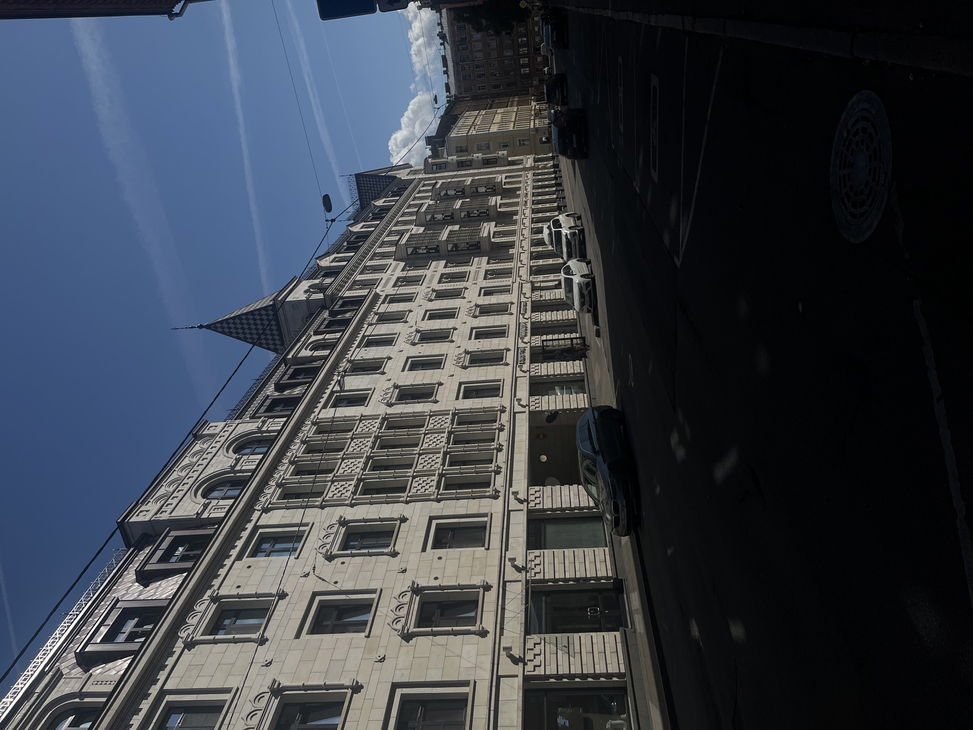 This is a medium shot, taken outdoors on a sunny day, of a city street in Saint Petersburg, Russia. The street is lined with ornate, multi-story buildings, featuring classical architecture with detailed facades, arched windows, and mansard roofs. Several cars are parked and driving on the street. The sky is blue with scattered white clouds and contrails from an aircraft. The perspective is from street level, looking down the street, with the buildings occupying the left side of the frame and the street and shadows dominating the right. The image captures a typical urban scene with architectural elements characteristic of Saint Petersburg.