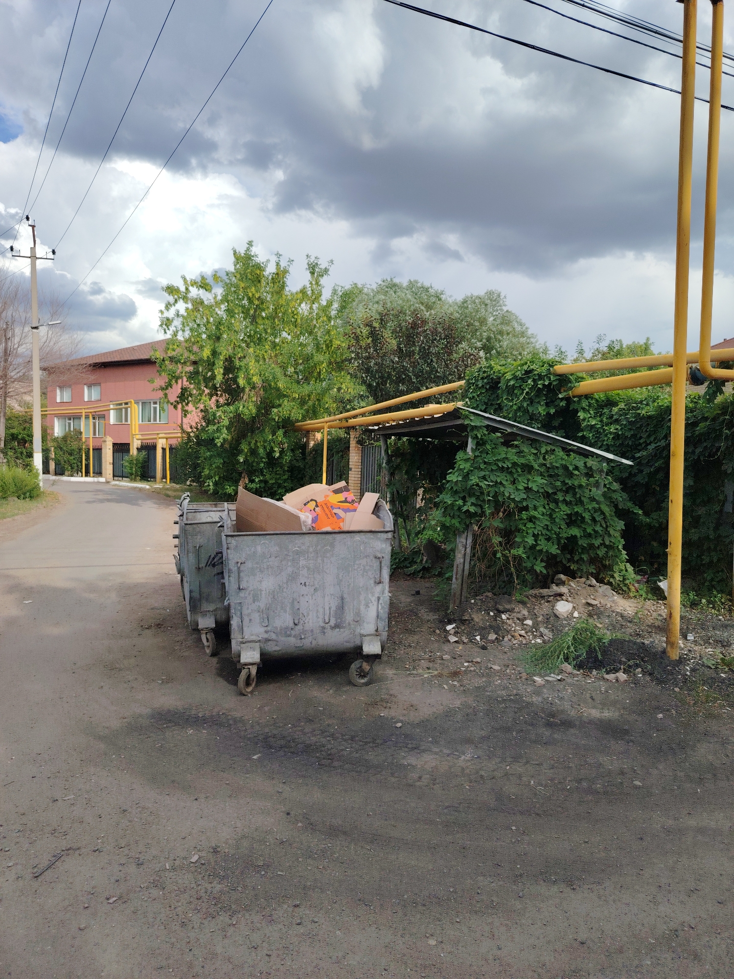 The image shows two large, weathered metal dumpster bins filled with cardboard and other debris. They are positioned on a dirt and gravel road, suggesting an outdoor, possibly semi-rural or industrial setting. In the background, a two-story building with a reddish-brown facade and white-trimmed windows is visible, with some green trees and bushes in front of it. Yellow pipes, possibly for gas or utilities, run horizontally along the building's facade and then vertically, forming a partial frame on the right side of the image. Further back, power lines stretch across a cloudy sky. The weather appears overcast, with dramatic, dark clouds suggesting an impending storm or recent rain. The overall scene is one of an ordinary day, with no people or animals present. The location is identified as Astana, Kazakhstan.