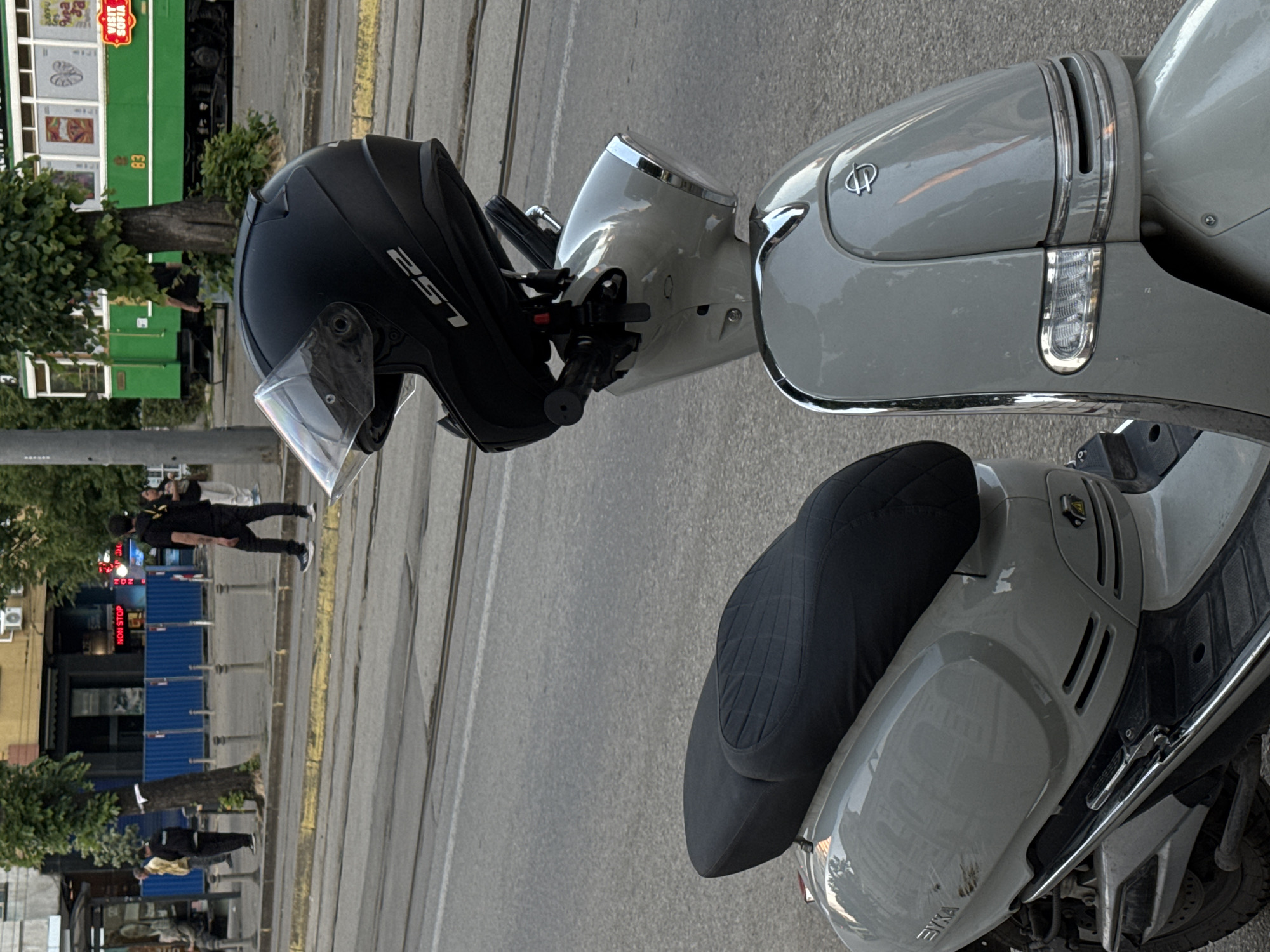 The image depicts a light gray Sym scooter parked on a city street in Sofia, Bulgaria. A black LS2 motorcycle helmet is mounted on the handlebars. Pedestrians walk on the sidewalk in the background, past businesses with blue and white signage. A green streetcar is visible in the background. The street appears to be paved with asphalt and includes tram tracks.