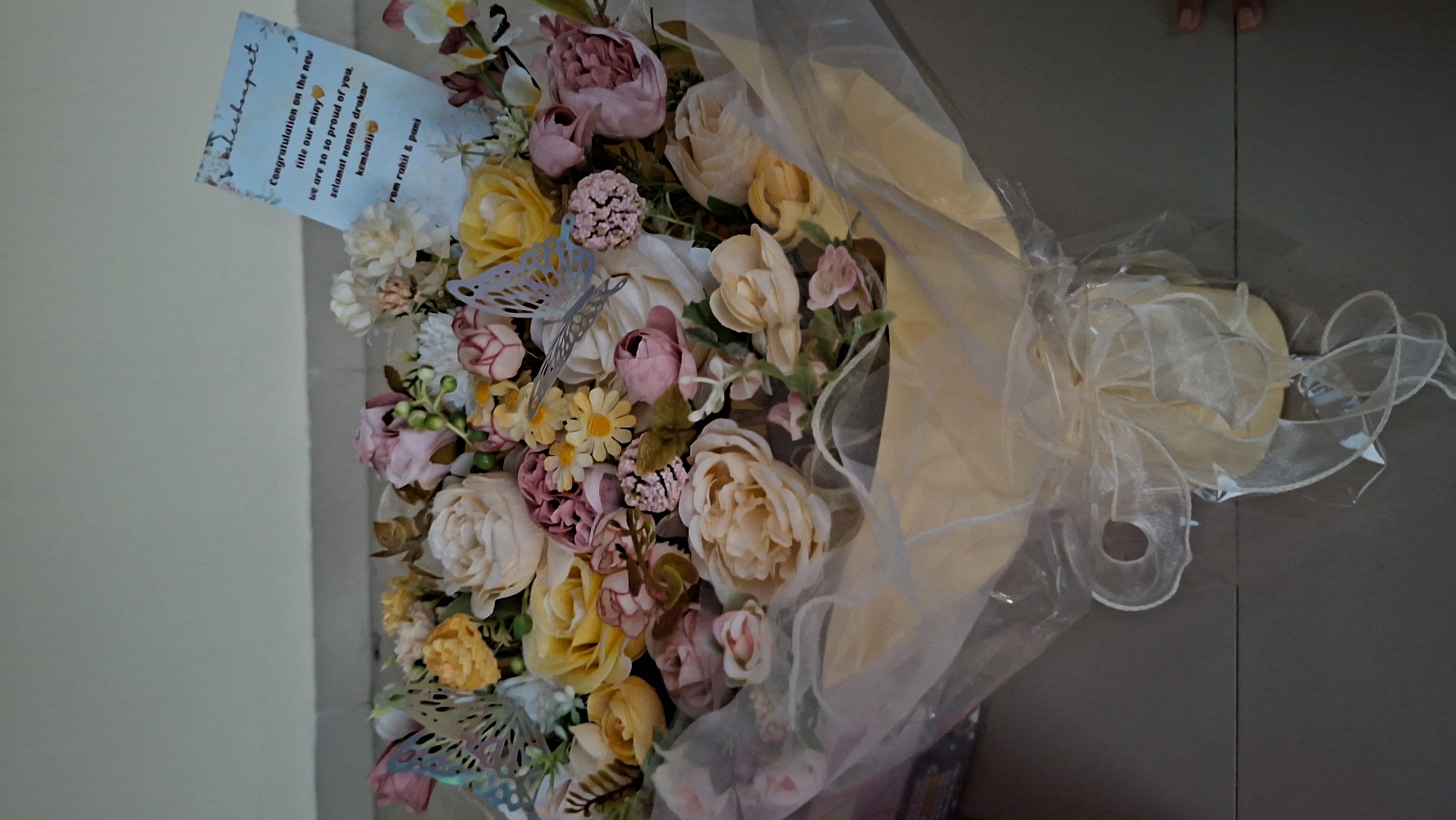 The image depicts a bouquet of artificial flowers, predominantly in pastel shades of pink, yellow, and cream, arranged with decorative butterflies. A congratulatory card is tucked into the bouquet, partially obscured by the flowers. The card reads: "Congratulation on the new title our miny 💛 we are so so proud of you. selamat nonton drakor kembali 💛 rem rahil & pani." The bouquet is wrapped in sheer white plastic and tied with a white ribbon. The setting appears to be an indoor environment, possibly a home, with a grey tiled floor and a light-colored wall in the background. The image does not contain any specific visual cues that would allow for pinpointing a location within Lhokseumawe, Indonesia. There are no visible individuals in the image.