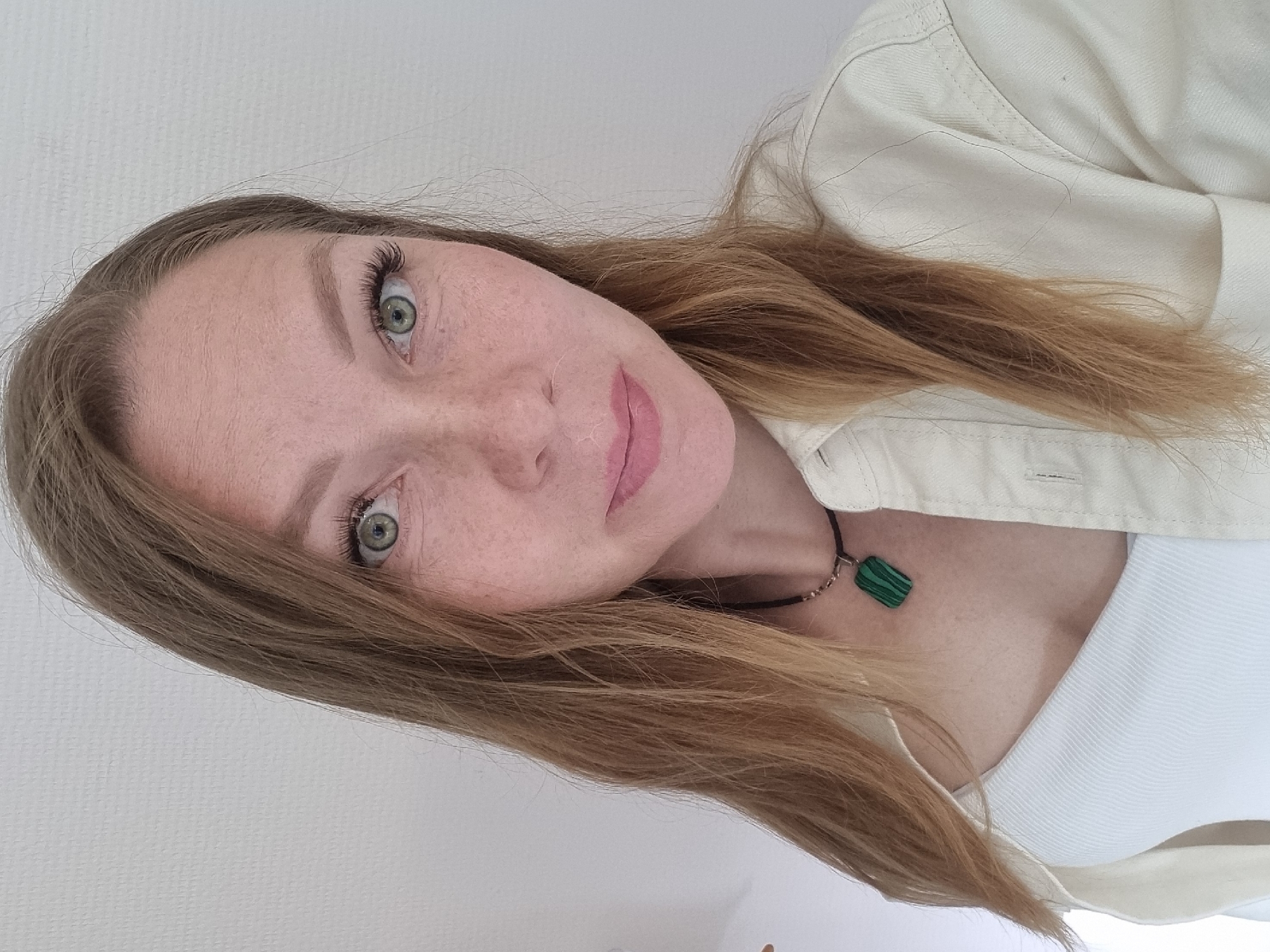 A medium shot shows a fair-skinned woman with long, light brown hair, a smattering of freckles, and green eyes. She is wearing a white ribbed tank top and an unbuttoned, off-white shirt. A dark cord necklace with a rectangular green pendant rests on her chest. The background is a plain white textured wall. The scene is indoors.