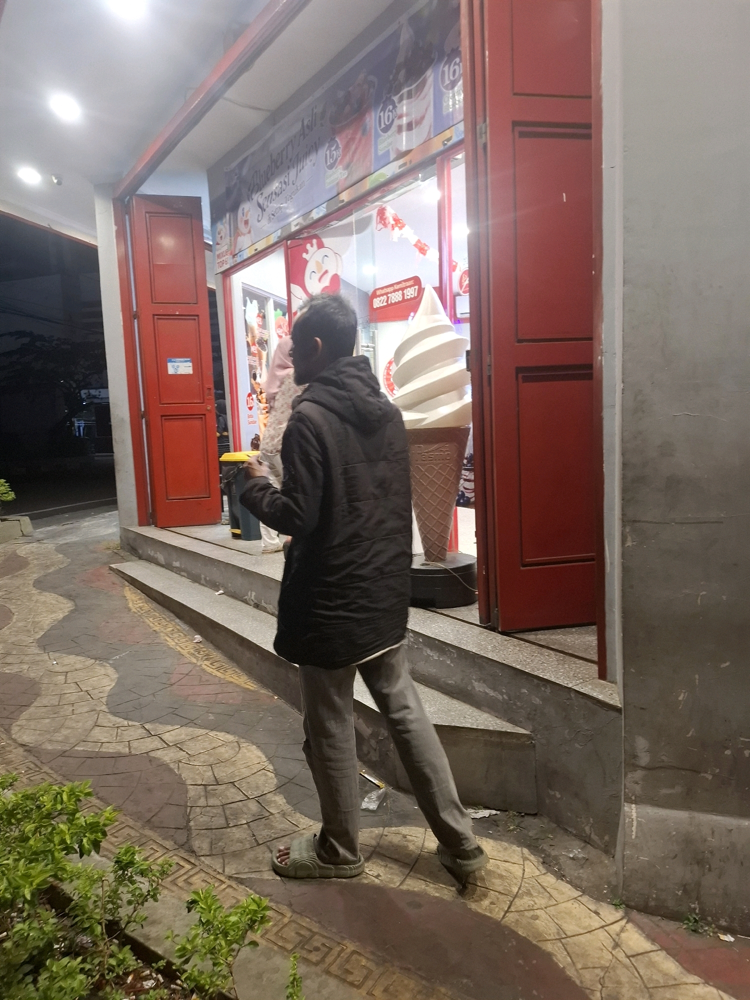 The image captures a street view in Cianjur, Indonesia, likely in the evening given the artificial lighting. A man in a black hooded jacket and grey pants is walking away from the viewer, towards an ice cream shop. The shop's entrance features large red doors and a display window showcasing ice cream cones, including a giant inflatable soft-serve cone. The pavement outside the shop has a decorative pattern of interlocking shapes and colors. There's a small green bush in the lower left corner of the frame, and a trash can is visible near the shop entrance. The overall environment suggests a commercial area with a somewhat worn appearance.