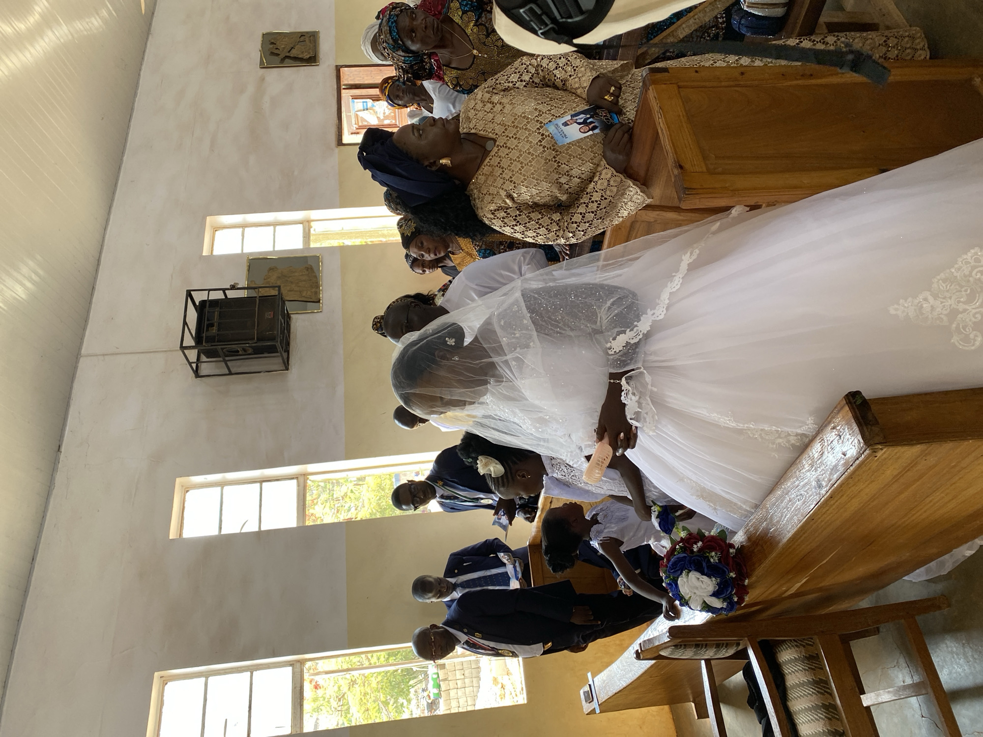 A wedding ceremony is taking place inside a church in Kuru Dungus, Nigeria. The bride, wearing a white wedding dress and veil, stands near the altar, where two young girls in white dresses are also present. Several men in dark suits with what appear to be ceremonial sashes or medals are positioned behind the girls. Seated in pews nearby are several women dressed in traditional African attire. The church's interior features cream-colored walls with windows letting in daylight, and wooden pews. Artwork and an air conditioning unit are visible on the walls.