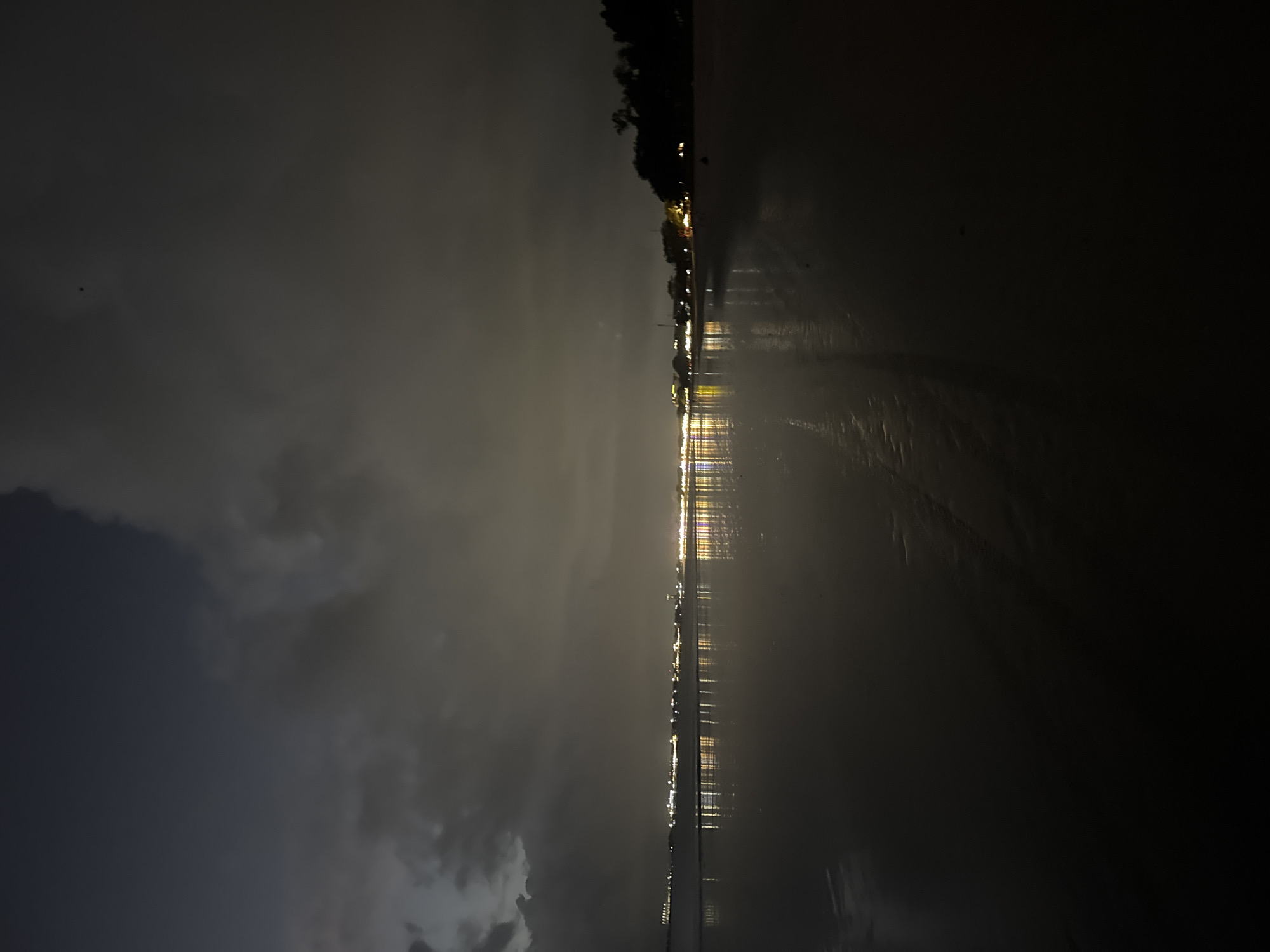 The image depicts a nighttime view of Kuta Selatan, Indonesia, likely from a beach. A dark, wet sand beach stretches into the foreground, reflecting the lights of the town. In the distance, a line of buildings with illuminated windows and lights are visible along the coast, creating a soft glow that is mirrored on the wet sand. The sky is dark and cloudy, with some lighter areas suggesting the presence of overcast skies or distant city lights. The overall impression is one of a quiet, atmospheric night scene. There are no people or animals clearly visible in the image.