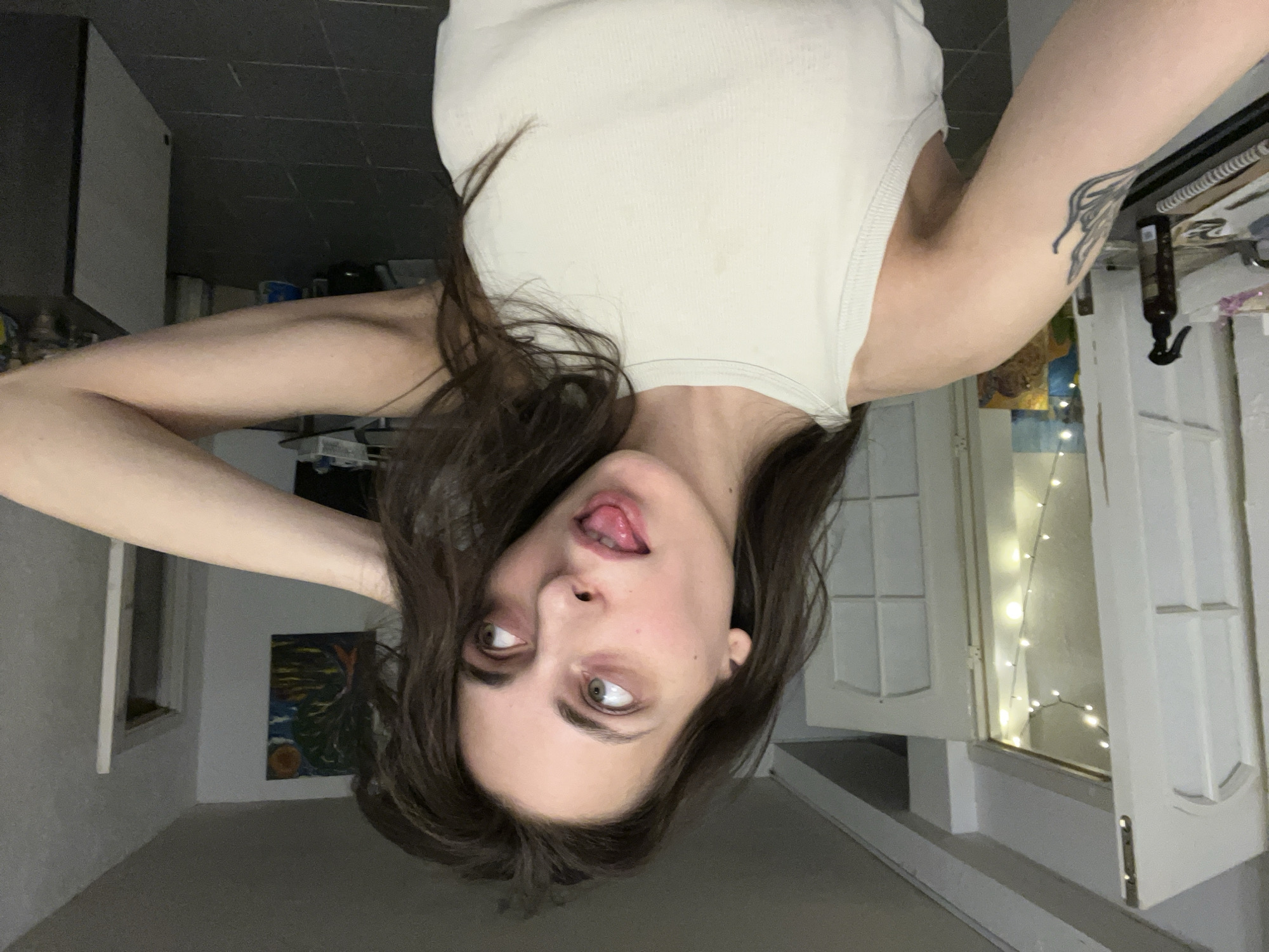 This image is an upside-down selfie of a person, likely female, with long, dark brown hair cascading around her face. She is wearing a light beige or off-white ribbed tank top. Her greenish-hazel eyes are rolled upwards, and her tongue is slightly out, conveying a playful or silly expression. A dark, possibly wing-like or abstract tattoo is visible on her right upper arm.
The setting is an indoor room, potentially in Krasnoyarsk, Russia, characterized by light grey walls and a darker ceiling. To the right, a white-framed window with small, warm fairy lights is visible, suggesting evening or night, or purely decorative lighting. Another window or opening is seen further back on the left. A small, colorful abstract painting hangs on a distant wall. The background also shows indistinct furniture or shelves. The overall mood is candid and informal. There is no visible text in the image.