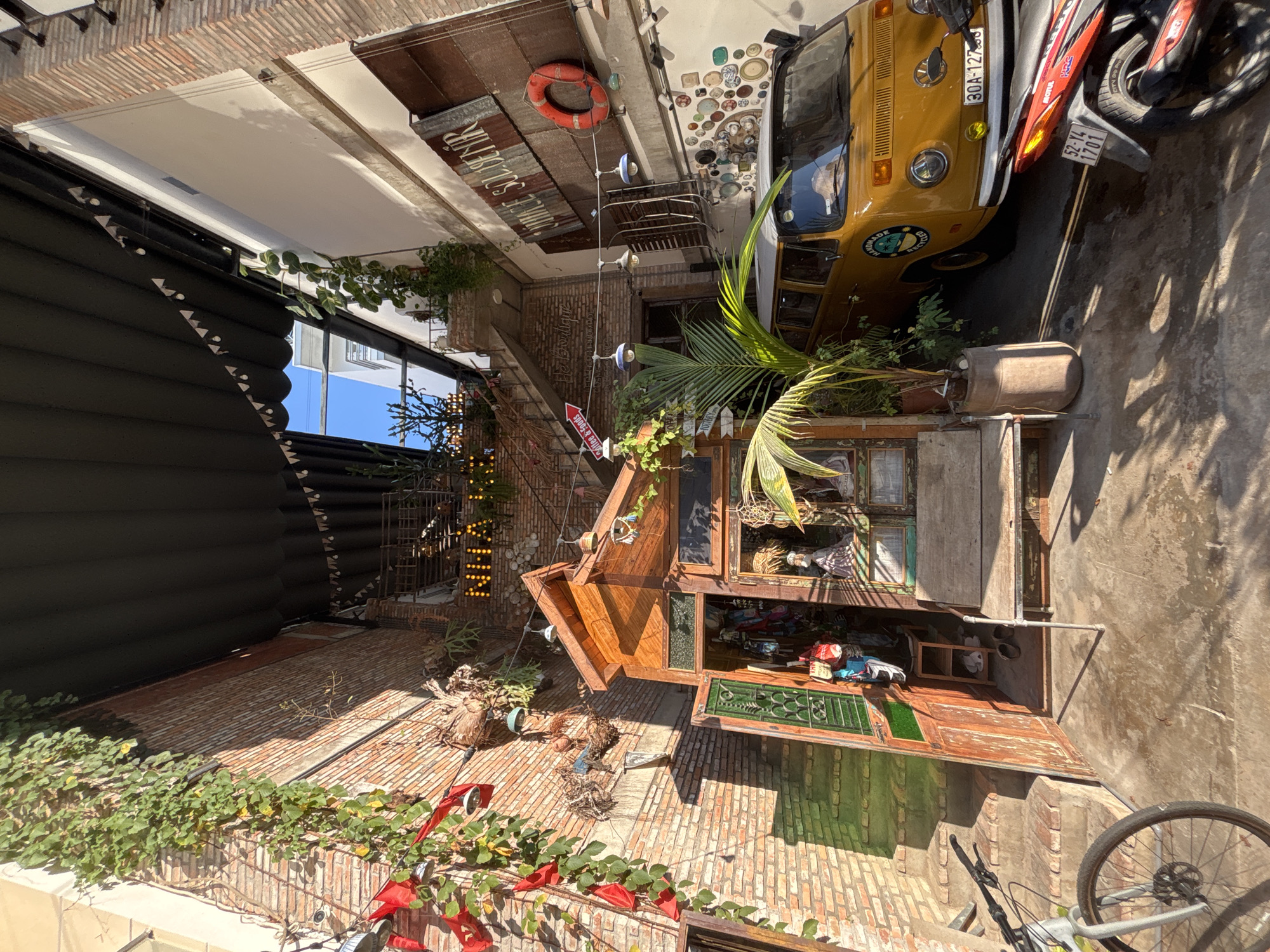 The image depicts an outdoor alleyway or courtyard in Nha Trang Ward, Vietnam, with a distinct bohemian and vintage aesthetic. The scene is dominated by weathered brick walls and a dark, corrugated awning stretched overhead. A small, rustic wooden stall or shop is a focal point, with its doors open, revealing an interior filled with various items, possibly handicrafts or apparel, hanging on display. Green-tinted glass is visible in the stall's windows and doors.
Adjacent to the stall, a weathered wooden bench sits outside. A bright yellow vintage Volkswagen van is parked to the right, adorned with a circular decal depicting a tropical scene and the words "HANDMADE" and "RECYCLED." The van has a license plate reading "30A-127.05". In the foreground, the front wheel and handlebars of a bicycle are visible on the left. A red and black motorcycle is parked to the right of the van, its license plate reading "52-Y4 1701".
Decorative elements include red fabric banners hanging from the wall on the left, string lights with small triangular flags, and various hanging ornaments. Plants, including a large palm, are integrated into the scene, adding to the natural and somewhat overgrown feel. A sign with red lettering spelling out "NHÀ HÀNG" (restaurant) is illuminated and mounted on a brick wall further back, suggesting a food establishment is also present. The ground is paved with concrete, showing signs of wear and some water pooling. The overall impression is one of a charming, eclectic space that blends retail, dining, and a relaxed atmosphere.