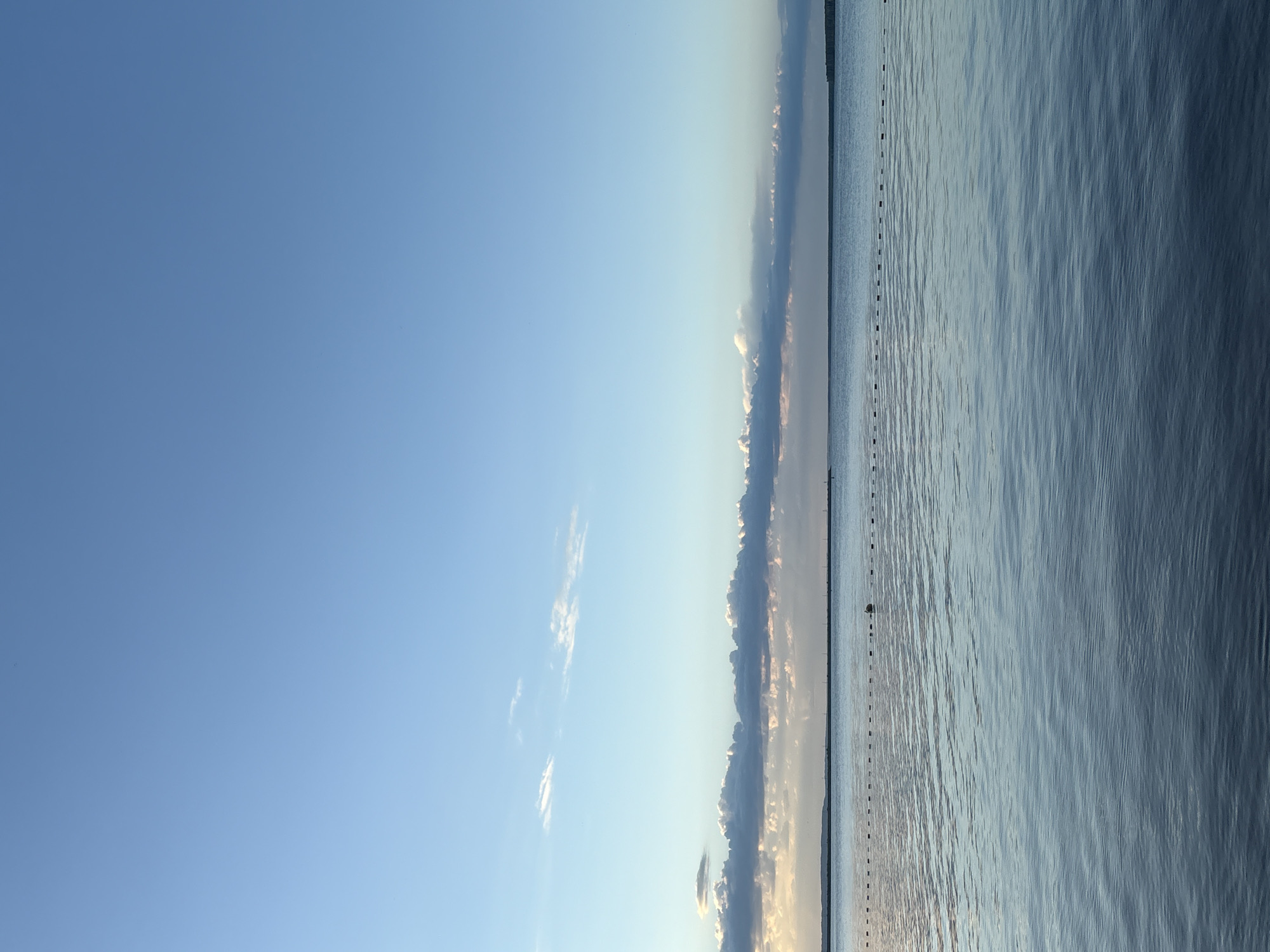 This image, taken in Chaikovsky, Russia, presents a tranquil waterscape under a vast sky, likely captured during early morning or late afternoon. The horizon line bisects the frame horizontally. The upper half is dominated by a clear, bright blue sky with a gradient of lighter tones near the horizon. Wispy white clouds are scattered across the higher sky, while a more substantial, elongated band of darker clouds stretches across the middle distance, exhibiting soft, warm light along its edges, indicative of either sunrise or sunset.
The lower half of the image features a calm body of water, presumably the Kama River or Reservoir, displaying subtle ripples across its surface and reflecting the soft light from the sky. A prominent line of small, dark, rectangular objects, appearing to be buoys or markers, extends horizontally across the water, marking a path or boundary. A slightly larger, singular dark object, possibly another buoy, is visible on the water's surface in the mid-right. A faint, dark strip of land or trees delineates the distant shoreline across the water. The scene is devoid of people or visible activities. No text is visible. The overall impression is one of peacefulness and natural serenity, under clear and calm weather conditions.