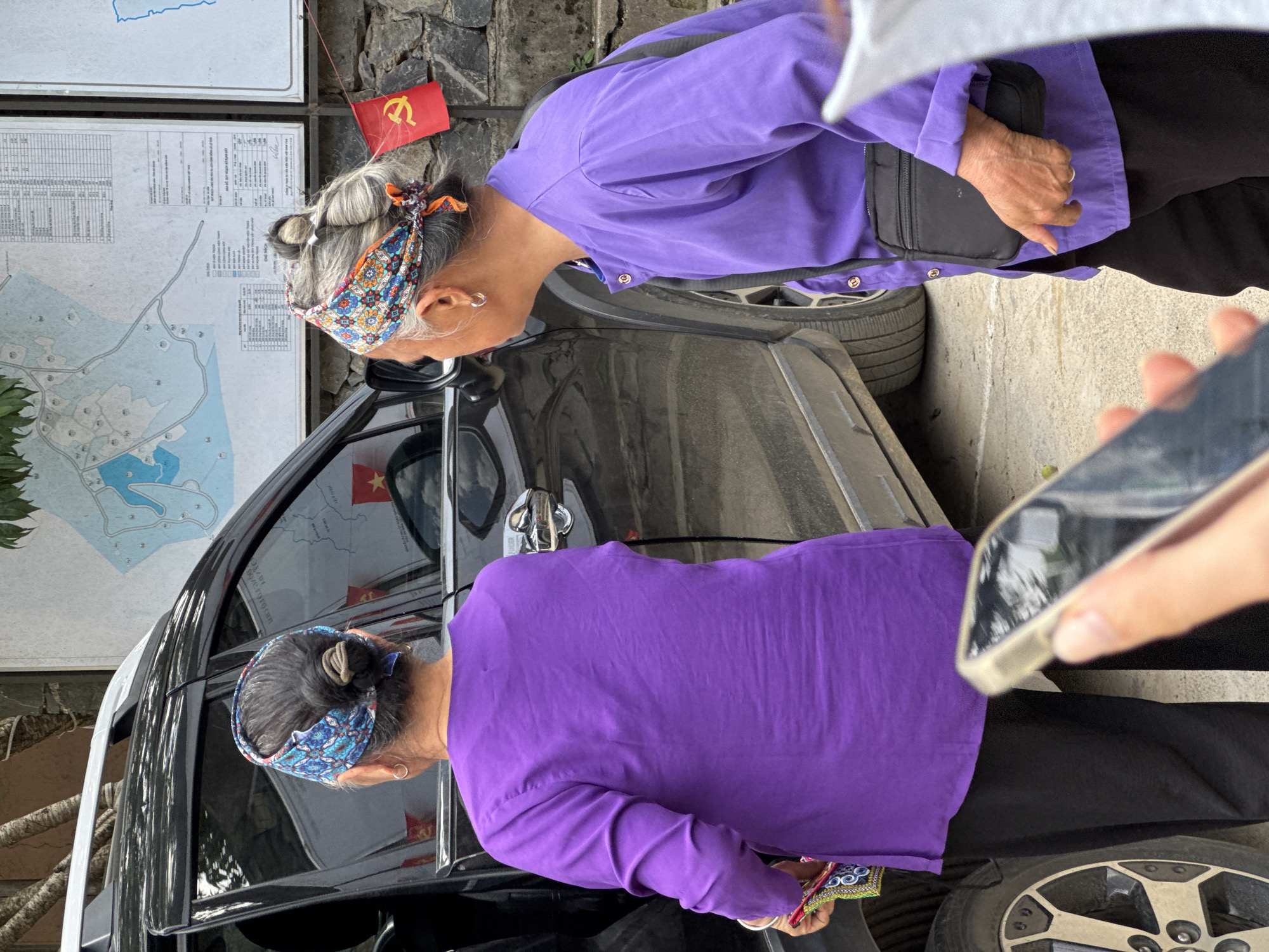 Two women are standing near a black car on the side of a street, with a map and information sign visible behind them. One woman, on the left, is closer to the car, looking towards the inside. She wears a blue patterned headscarf and a purple shirt. The other woman, to the right, also wears a purple shirt and black pants. Both appear to be in an urban environment. The presence of the flag suggests this is located in Vietnam, possibly related to the city of Lũng Cú.