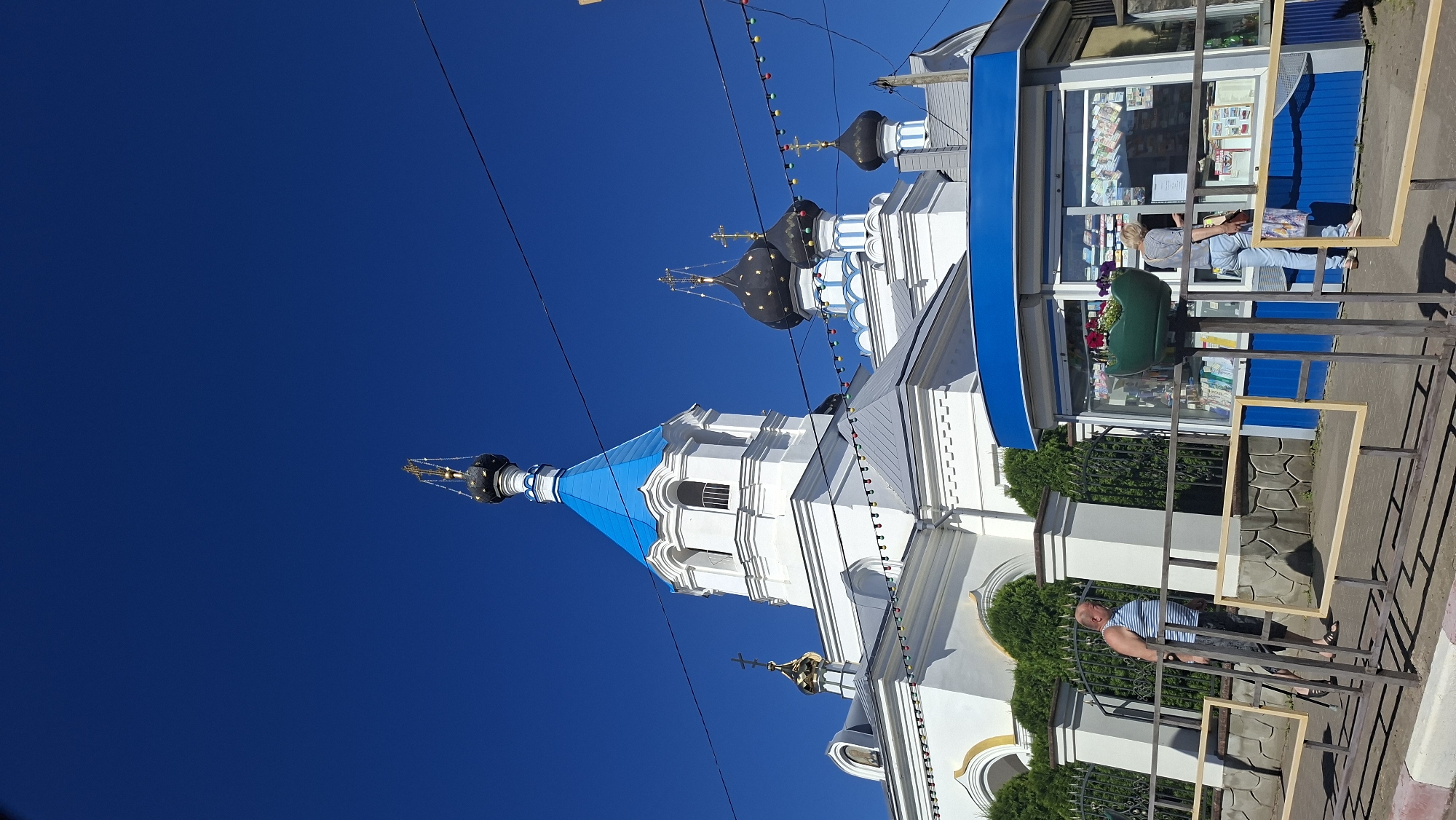 This is a full-shot, slightly upward-angled, vertical photograph of the Church of the Holy Trinity in Pastavy, Belarus, on a clear, sunny day. The church, a white building with blue-domed roofs and gold crosses, dominates the upper portion of the frame. In the foreground, a man in a striped tank top and shorts walks on a sidewalk next to a black metal fence. To the right of the fence is a blue and white kiosk with its windows displaying various items. A woman in light blue jeans and a striped shirt stands at the kiosk, appearing to be making a purchase. Several strings of colorful lights are strung across the scene, some hanging low, and power lines are visible against the bright blue sky. The area around the church is green with trees and bushes.