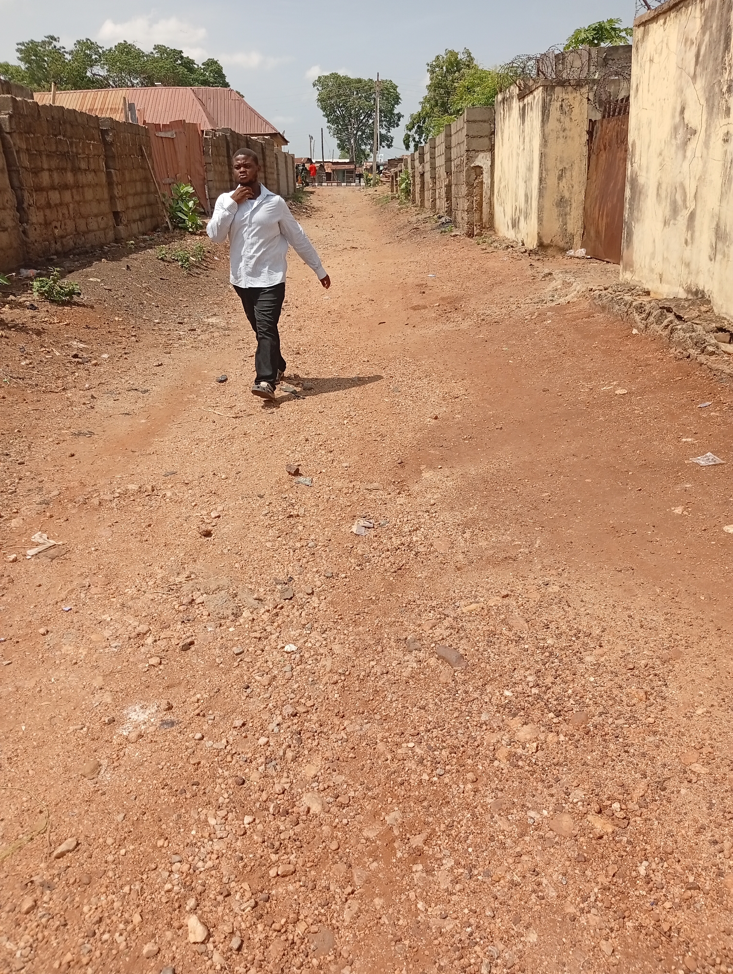 The image depicts a man walking on an unpaved road in Adankolo, Nigeria. The road is reddish-brown and covered with small stones and debris. The man is wearing a light-colored, button-down shirt and dark pants. He appears to be adjusting his collar. Walls made of concrete blocks or painted concrete structures line both sides of the road. Some of the walls have sections with barbed wire on top. In the distance, other people and structures are visible, including a building with a corrugated metal roof. The sky is partly cloudy.