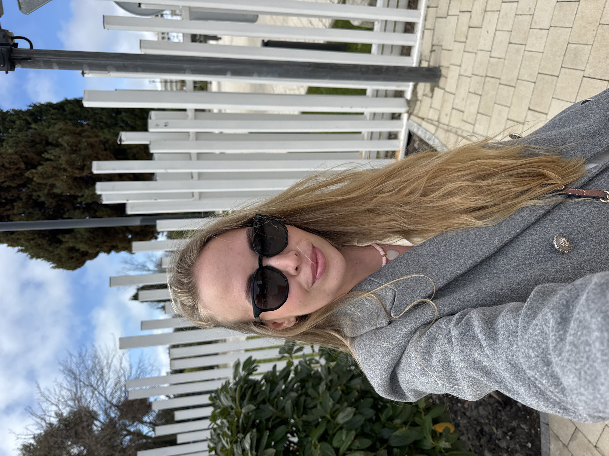 A woman with long blonde hair, wearing a gray coat and sunglasses, is taking a selfie outdoors. In the background, there is a white metal fence, greenery, and a brick sidewalk. The sky is visible in the upper left corner. Based on the architecture and vegetation, the location appears to be in the city of Sochi, Russia.