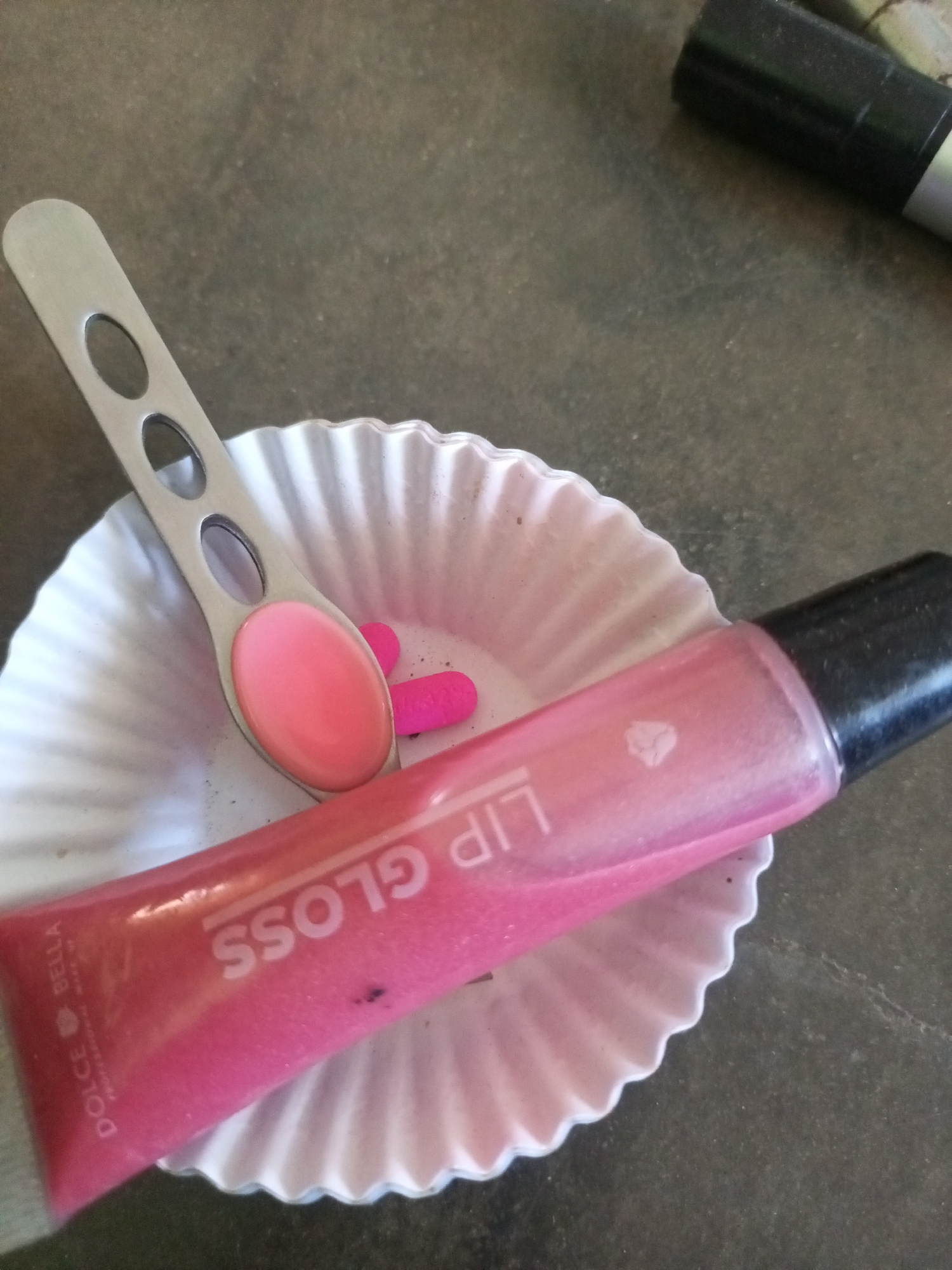 The image displays a close-up of a pink lip gloss tube, a small metallic spatula with a pink substance on it, and two small pink pills or capsules, all resting inside a fluted paper cup. The lip gloss tube has text that appears to read "LIP GLOSS" in white, large letters, and "DOLCE BELLA PROFESSIONAL MAKE UP" in smaller text on the side. The metallic spatula has two oval-shaped holes in its handle. The background is a dark, textured surface, possibly a table. The scene is a still life arrangement of makeup and what appear to be medication. There are no people or any context to suggest a specific location in La Tagua, Venezuela.