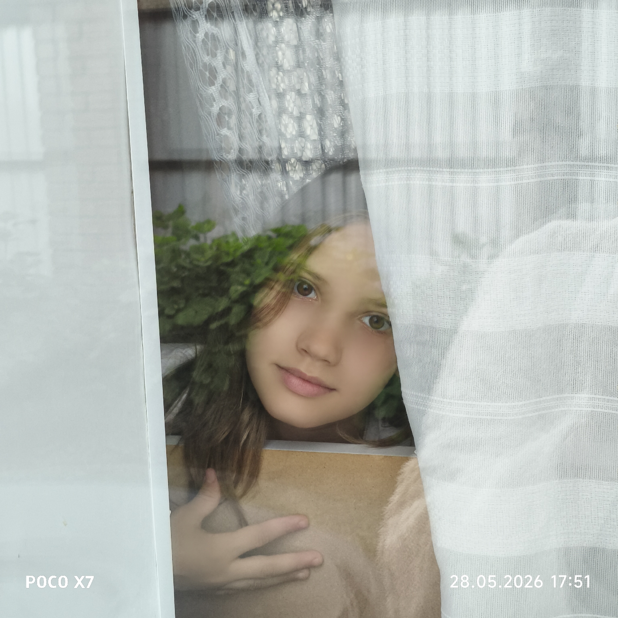 A girl is looking through a window covered by a curtain. The window has two layers of curtains, one with a lacy pattern and one that is vertically striped. Green plants are visible behind the girl. The girl is holding a book or a notebook. The date and time "28.05.2026 17:51" and the text "POCO X7" are printed on the image. The location is Unknown, Unknown.