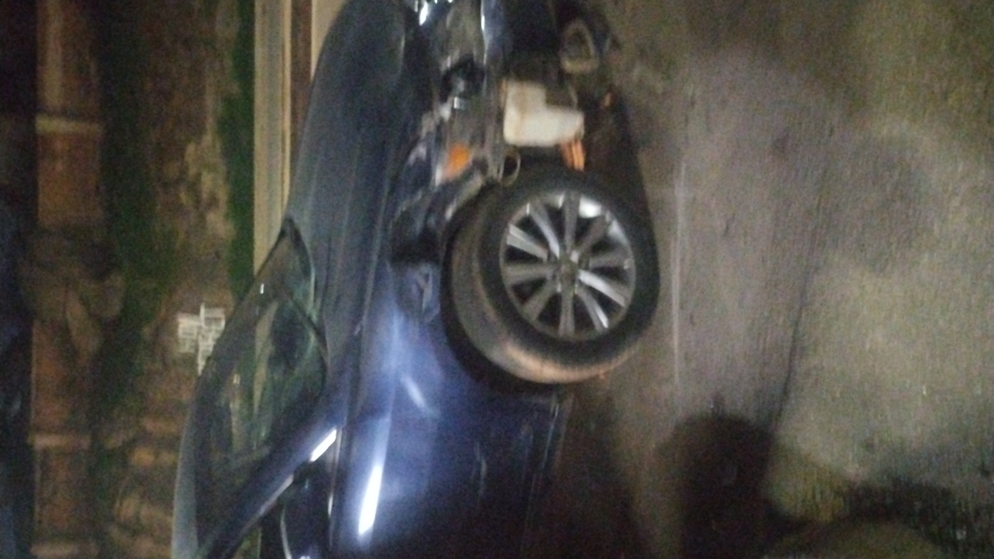 A dark-colored sedan is overturned and leaning against a concrete wall in an urban environment, possibly the city of Kabong, Nigeria. The vehicle's wheel is prominently visible, and its undercarriage is exposed. The surrounding area appears dimly lit.