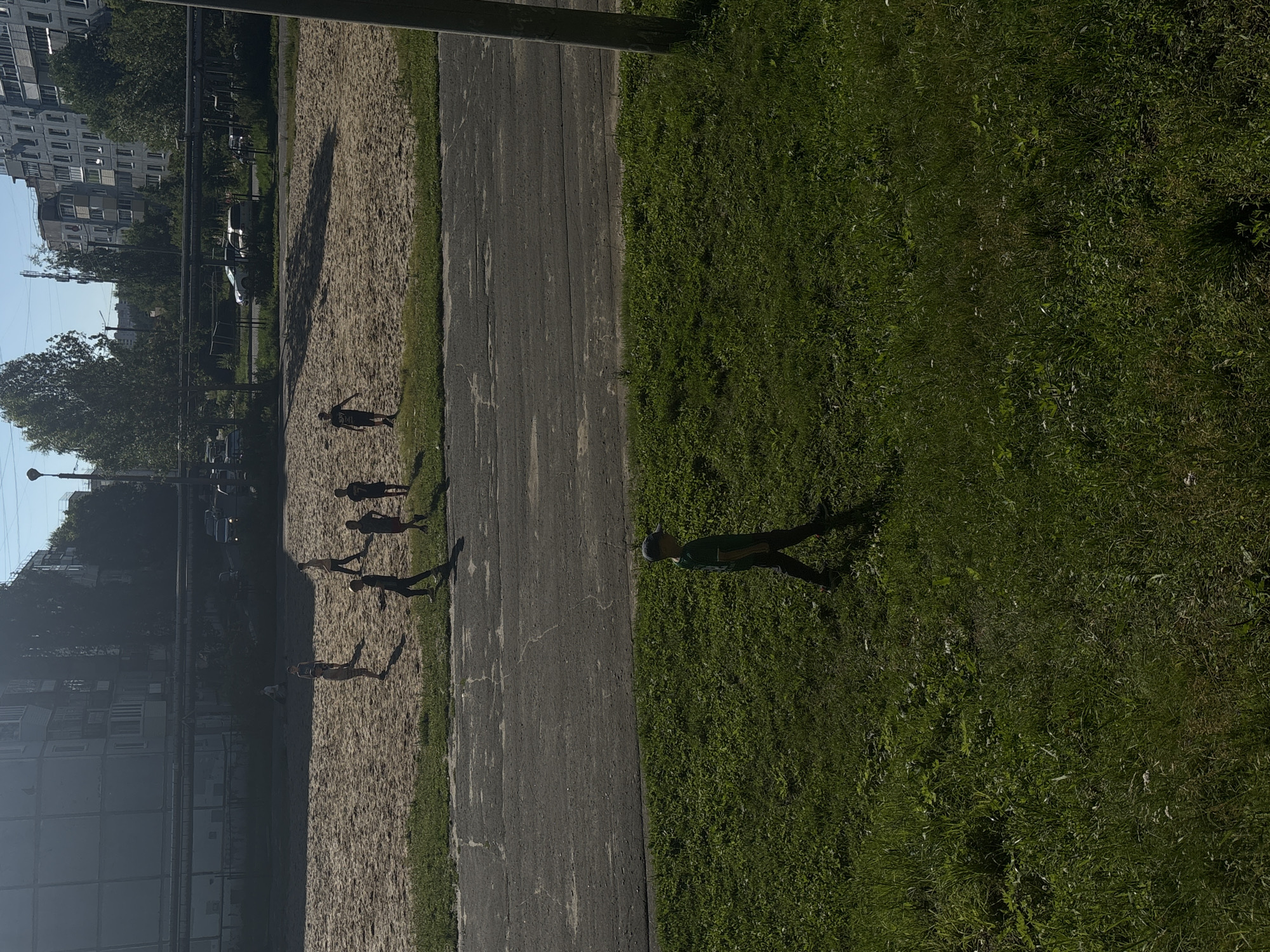 The image depicts a sunny outdoor scene in Yoshkar-Ola, Russia. In the foreground, a young boy wearing a green t-shirt and a dark baseball cap walks across a grassy area, casting a shadow. A paved road with visible cracks runs horizontally across the middle of the image. Beyond the road lies a sand-covered area, possibly a sports court, where several children are engaged in activity. Some of the children are standing, while others appear to be running or playing. In the background, apartment buildings and trees are visible under a clear blue sky. Several cars are parked on a road in the distance. The overall scene suggests a residential area with recreational space.