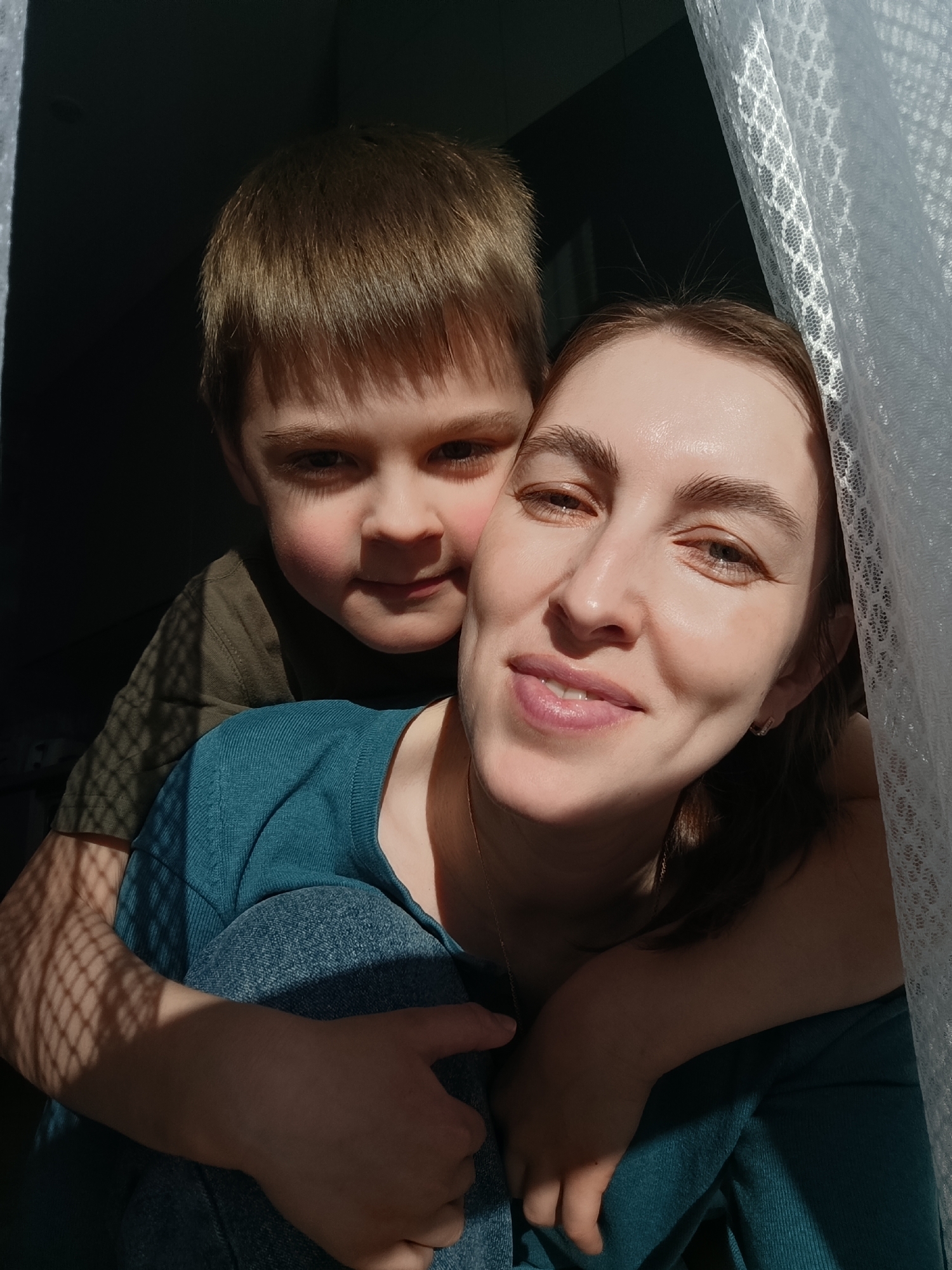 The image shows a woman and a young boy posing indoors. The boy is behind the woman, with his arms around her neck and shoulders. The woman is smiling and looking at the camera. They are close together, possibly embracing. They are near a window. The background is dark. The location is Chaikovsky, Russia.