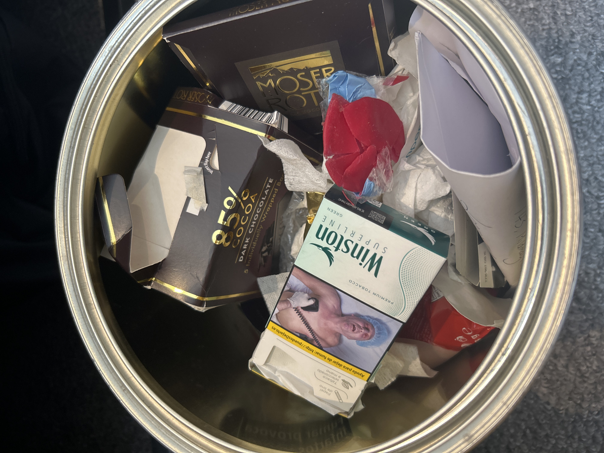 The image depicts a metallic trash can filled with various discarded items. Among the recognizable objects are several chocolate wrappers from "Moser Roth" and a cigarette pack of "Winston Superline GREEN" brand. There are also pieces of crumpled paper and packaging materials mixed in. The trash can is located in an unspecified indoor environment. There is no indication of it being located in London, United Kingdom.