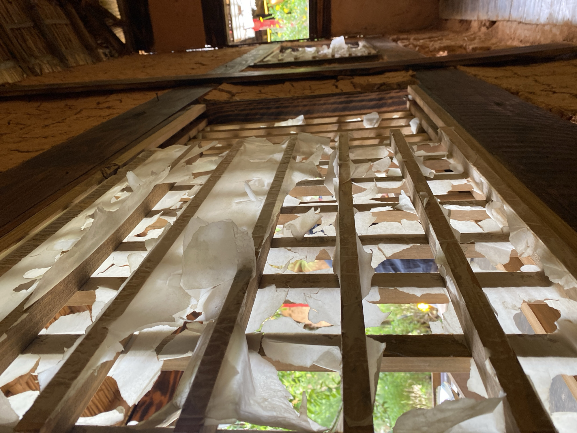 The image shows a close-up view of a traditional wooden lattice window in Da Lat, Vietnam, with torn, translucent paper applied to it. The paper is partially covering the wooden slats, creating a textured and fragmented effect. Through the openings in the lattice and torn paper, glimpses of lush green foliage and a glimpse of a person in the background are visible. The interior of the building features rough, earthen walls and a wooden ceiling structure. The lighting suggests an outdoor environment.