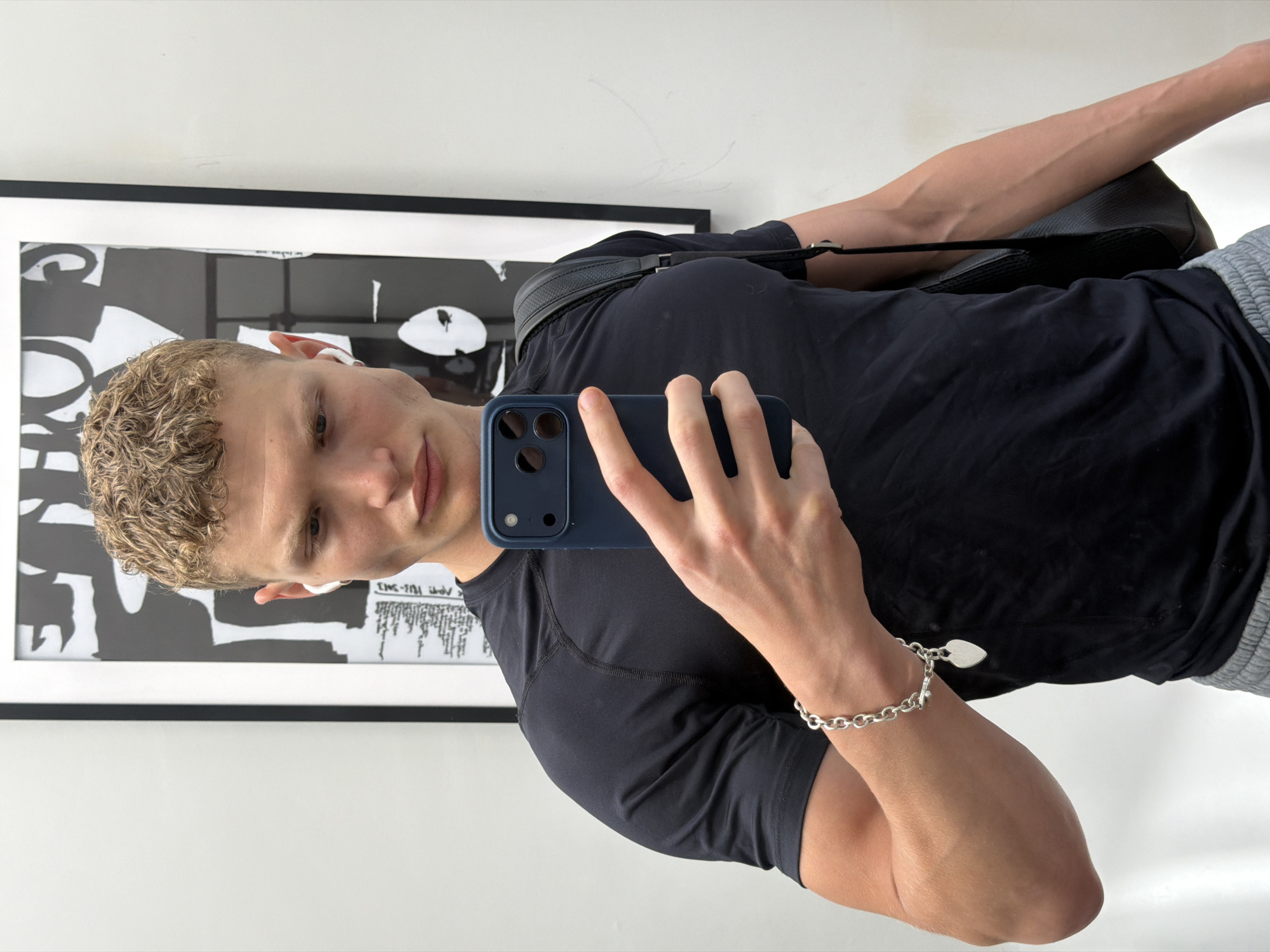 A young man with short, curly blonde hair, wearing a black t-shirt, grey sweatpants, white earbuds, and a silver bracelet with a heart charm, stands in front of a mirror, taking a selfie with a dark blue smartphone. He is also wearing a black bag over his left shoulder. Behind him, a framed black and white abstract print is visible on the wall, as well as a white wall. The scene appears to be an indoor environment, possibly a changing room or a home, with no specific indicators of a London location.