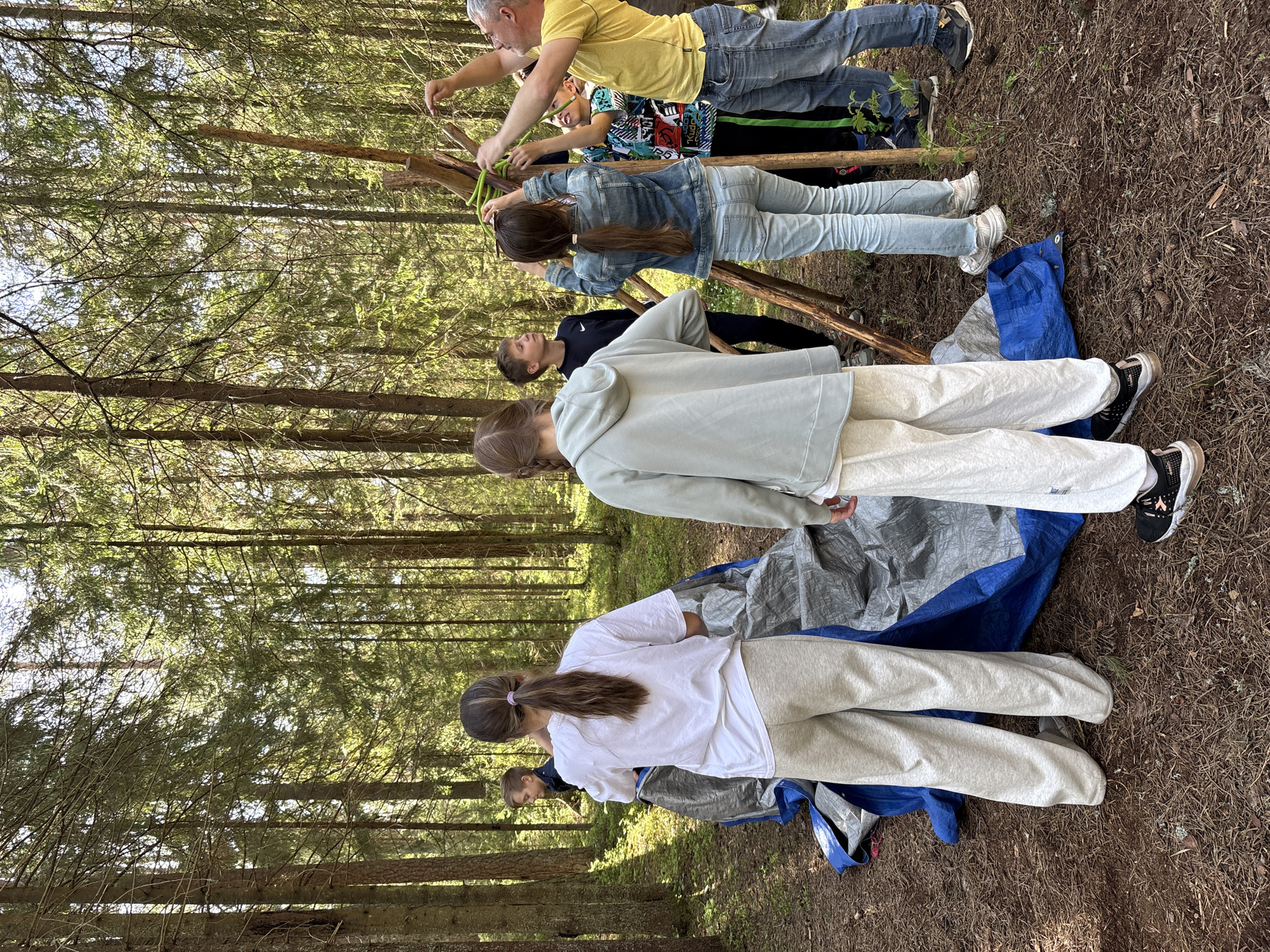 The scene is an outdoor setting within a forest, likely during daylight hours due to the visible sunlight filtering through the trees. The ground is covered with pine needles and other natural debris.
A group of people, including what appears to be a mix of adults and children, are engaged in a construction activity. They are using wooden sticks and what looks like rope or cord to create some form of structure. A tarp, composed of both blue and gray sections, lies on the ground beneath them.
One adult is wearing a yellow shirt and jeans. Other individuals are dressed in casual clothing like T-shirts, sweatpants, and denim jackets. They appear to be focused on building the structure.
The location is Unknown, Russia.