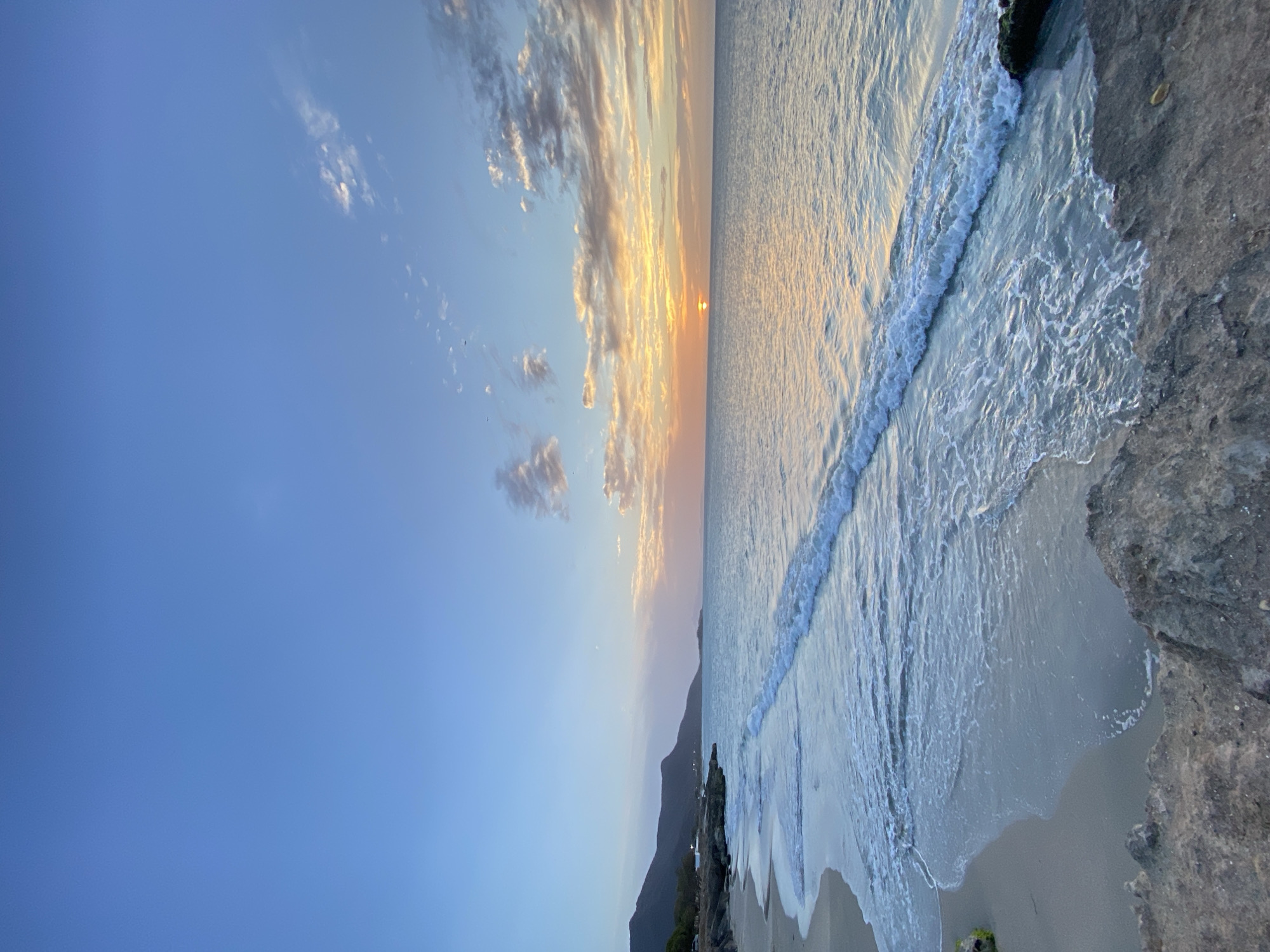 The image depicts a sunset view of the sea meeting the shore. The scene includes a view of the beach and the ocean with waves gently lapping the shore. In the distance, there's a mountain range, and the sky is partially covered with clouds illuminated by the setting sun. The scene is on Playa Caribe, Juan Griego, Venezuela.