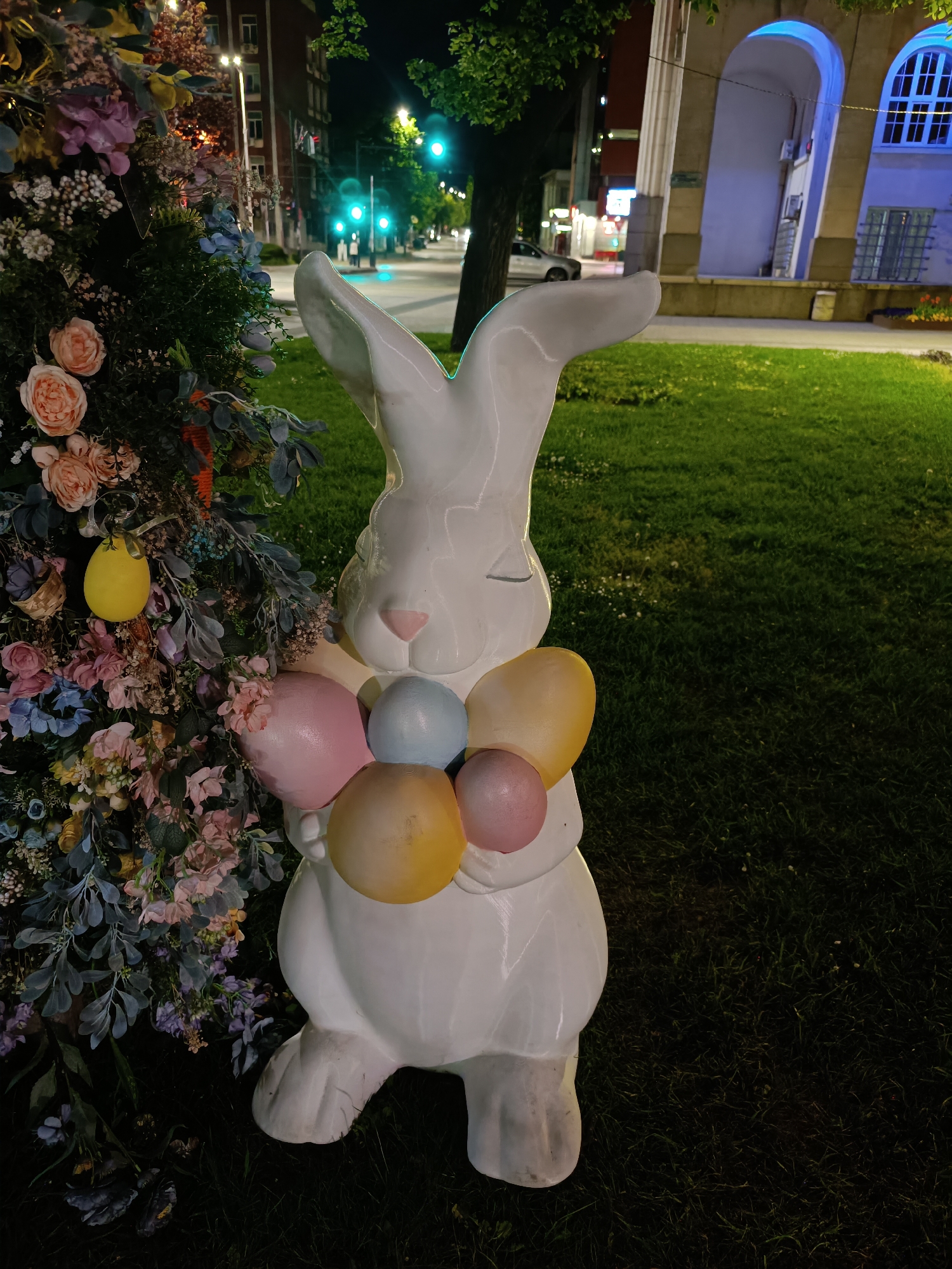 The image shows a white ceramic rabbit statue standing on a grassy lawn. The rabbit is decorated with pastel-colored, egg-shaped objects resembling balloons around its neck. To the rabbit's left, there's a colorful floral arrangement featuring pink, peach, and blue flowers. In the background, there's a street with streetlights, buildings, and cars, suggesting an urban environment. One building in the background is lit with blue lights in its arched windows. The scene appears to be at night. The location is Unknown, Unknown.