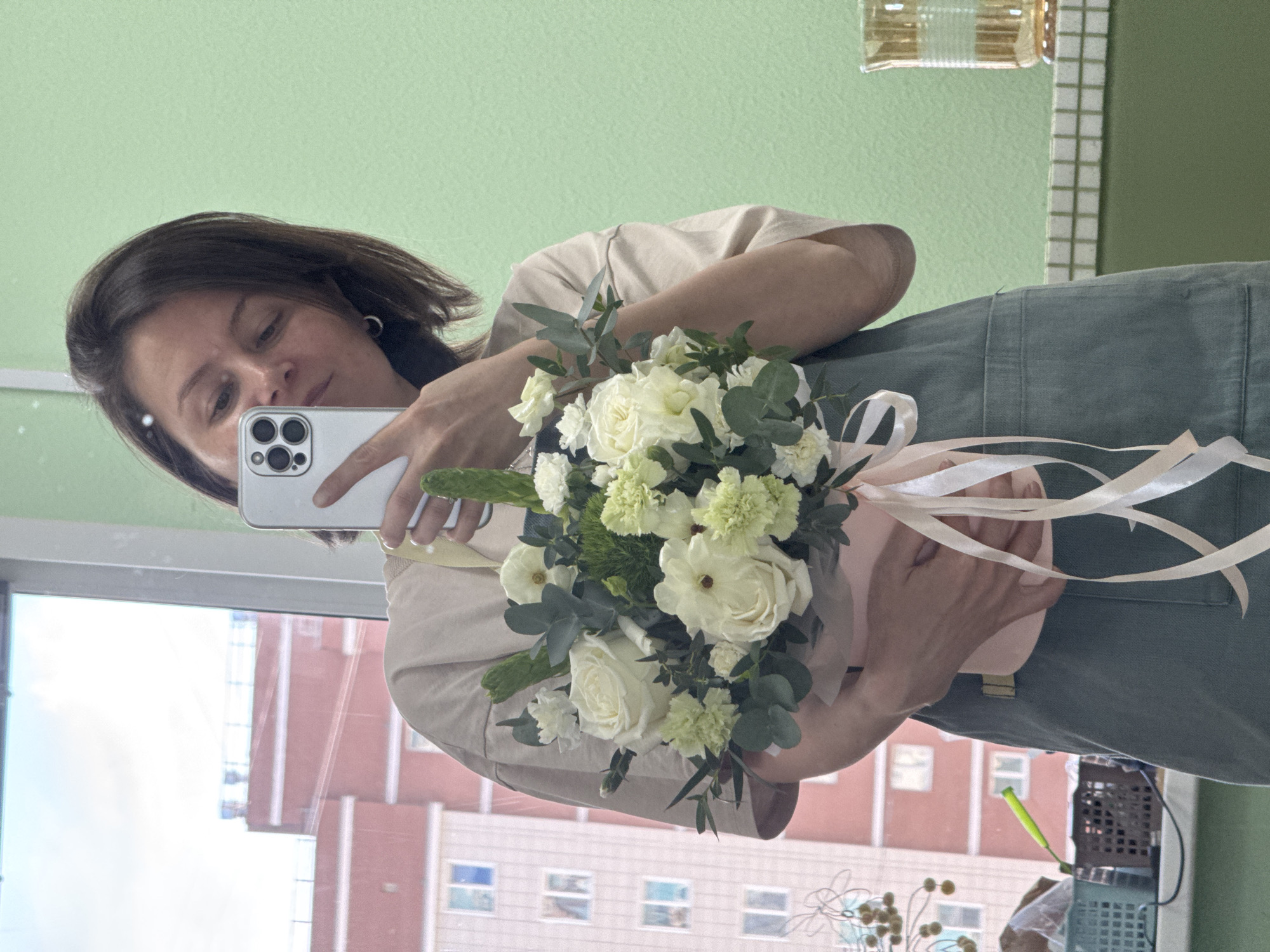 A woman holds a bouquet of white flowers while taking a selfie in a mirror. The background reveals a building outside the window, suggesting the location is likely Izhevsk, Russia, as previously mentioned. The woman wears a neutral-toned top and green overalls. The bouquet is wrapped in a light pink material and adorned with cream-colored ribbons. The mirror reflects a green wall with white tile detailing along the top. A bottle sits on the shelf above the tiles.