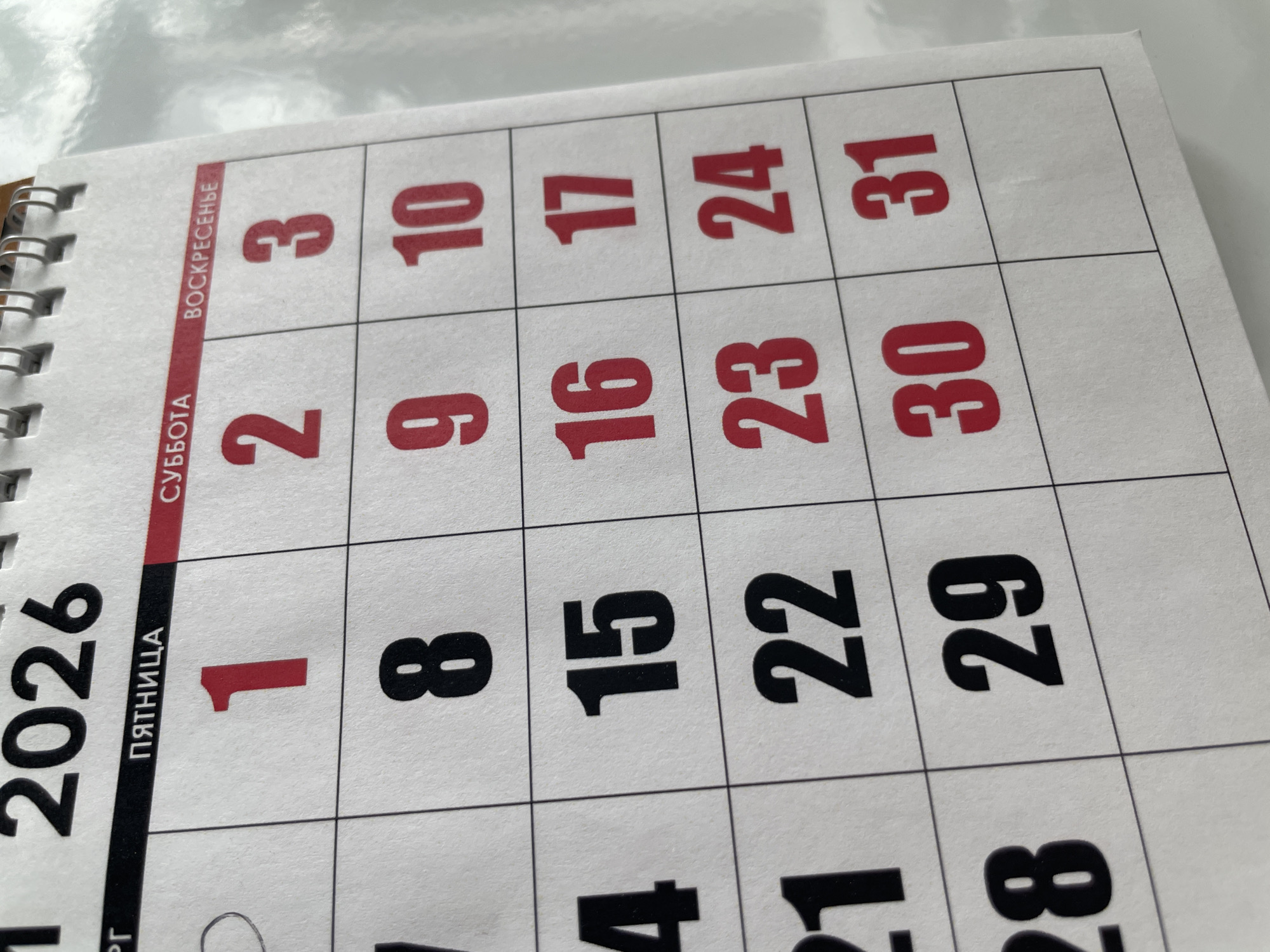 The image shows a close-up of a calendar for the year 2026. The top of the calendar displays the days of the week in Russian, with Friday ("ПЯТНИЦА") being the first day shown, followed by Saturday ("СУББОТА") and Sunday ("ВОСКРЕСЕНЬЕ"). The numbers 1 through 31 are arranged in a grid, representing the days of the month. Some of the dates are printed in red, while others are in black. The calendar is bound by a metal spiral on the left side.