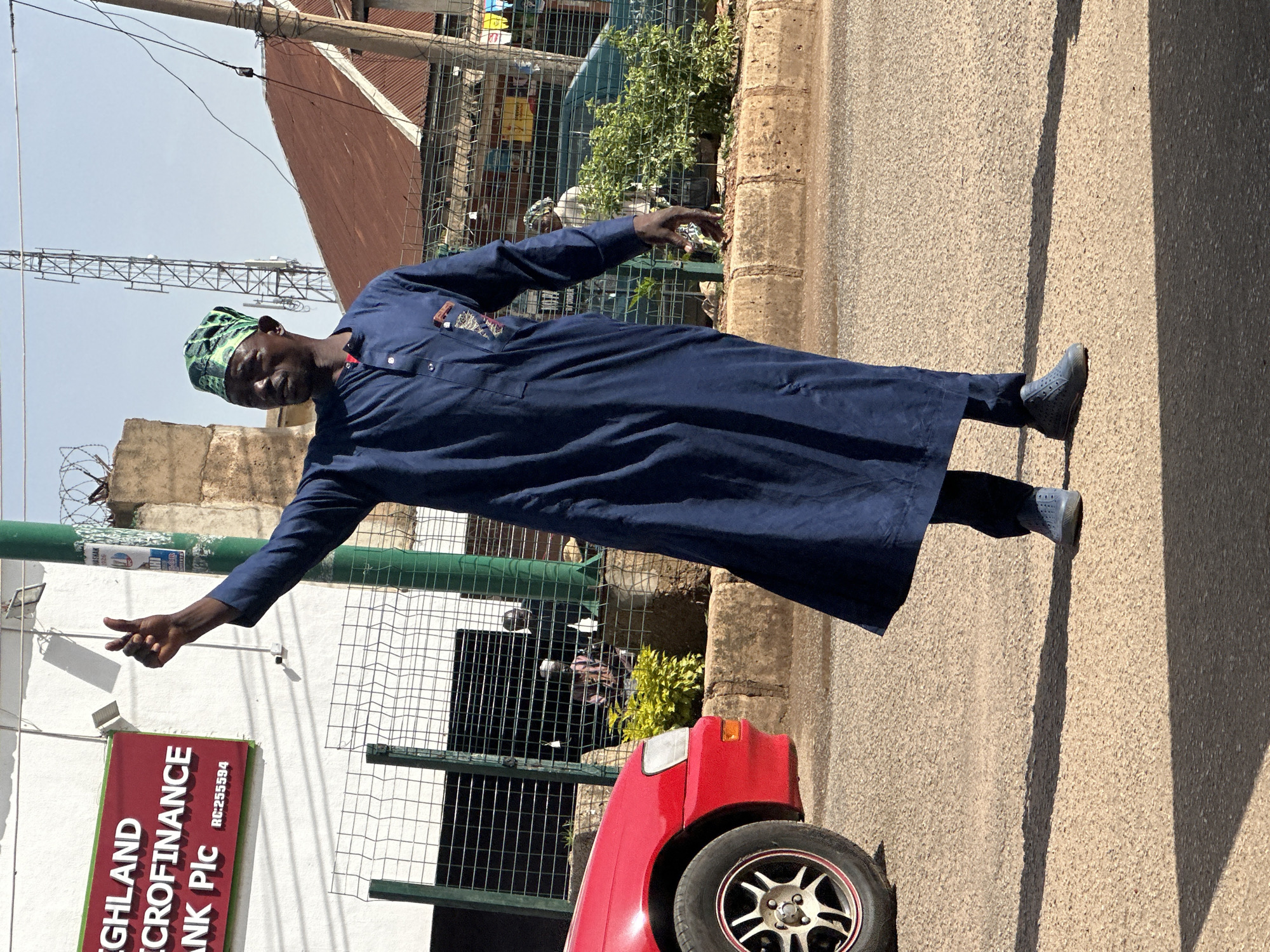 A man wearing a navy blue traditional outfit and a green and black patterned headwrap stands on a paved road in Bukuru, Nigeria. He is facing to his left and has his right arm raised with his thumb up, signaling. A red car is parked to his left, partially in the frame. Behind the man, a white building with a red sign that reads "HIGHLAND MICROFINANCE BANK Plc RC:255594" is visible. A green metal fence separates the sidewalk from the road. In the background, there are other buildings, a cell phone tower, and vegetation. The lighting suggests it is daytime with sunlight casting shadows on the road.
