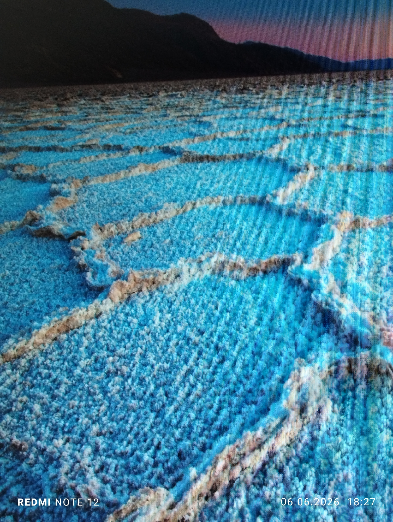 This image captures a wide, low-angle perspective of an outdoor landscape, attributed to the city of Burgas, Bulgaria. The foreground and midground are covered by an expansive, patterned surface resembling polygonal structures, likely a salt flat or frozen ground. These formations are characterized by a vibrant cyan-blue infill, sharply delineated by raised, irregular ridges composed of white, off-white, and light brown granular material, suggesting crystalline or rocky texture. The patterns diminish in size and clarity towards the horizon due to perspective. In the background, a dark, silhouetted mountain range spans the width of the frame. Above the mountains, the sky displays a twilight gradient, transitioning from deep indigo at the top to shades of purple and orange-pink along the horizon line. No individuals, animals, or man-made objects other than the geological features are present. The image exhibits significant visual artifacts, including prominent vertical scan lines and pixelation across the entire frame, indicating it is a photograph taken of a digital display. Overlaid text in the bottom left corner reads "REDMI NOTE 12," and a timestamp in the bottom right corner shows "06.06.2026 18:27."