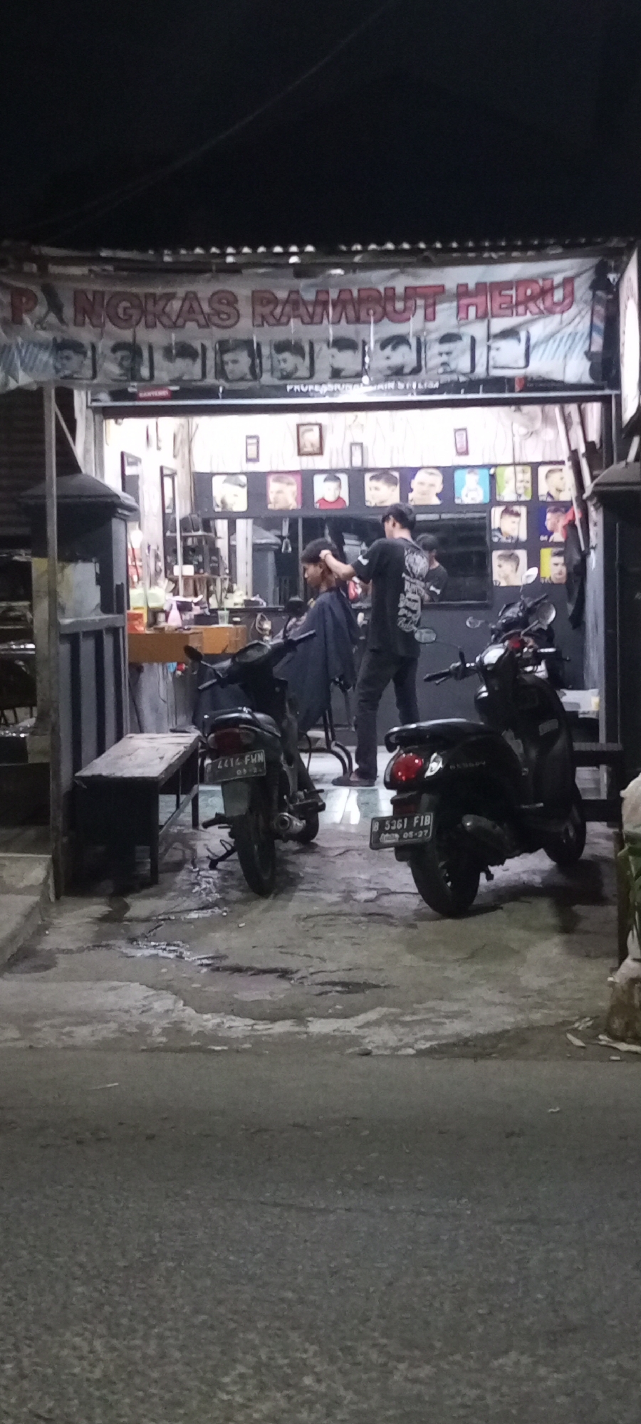 The image depicts a street-level view of a barbershop in Setu, Indonesia. The barbershop has a banner across the top with text that appears to read "PANGKAS RAMBUT HERU," indicating it is a hair salon named Heru. Below the banner, a series of framed pictures of hairstyles are displayed.
Inside the barbershop, two men are visible. One man, wearing a dark t-shirt with a graphic print on the back, is standing and appears to be cutting the hair of another person seated in a barber chair. The person in the chair is wearing a dark cape. Shelves with various items, possibly hair products and equipment, are visible behind them.
In front of the barbershop, two motorbikes are parked. The motorbike on the left has a license plate that reads "B 4414 FWM." The motorbike on the right has a license plate that reads "B 5361 FIB." The ground in front of the shop is wet and uneven, suggesting it may have recently rained or is a drainage area. The overall scene is lit by artificial lights, indicating it is likely nighttime. The environment appears to be a simple, open-air storefront typical of local businesses in Indonesia.