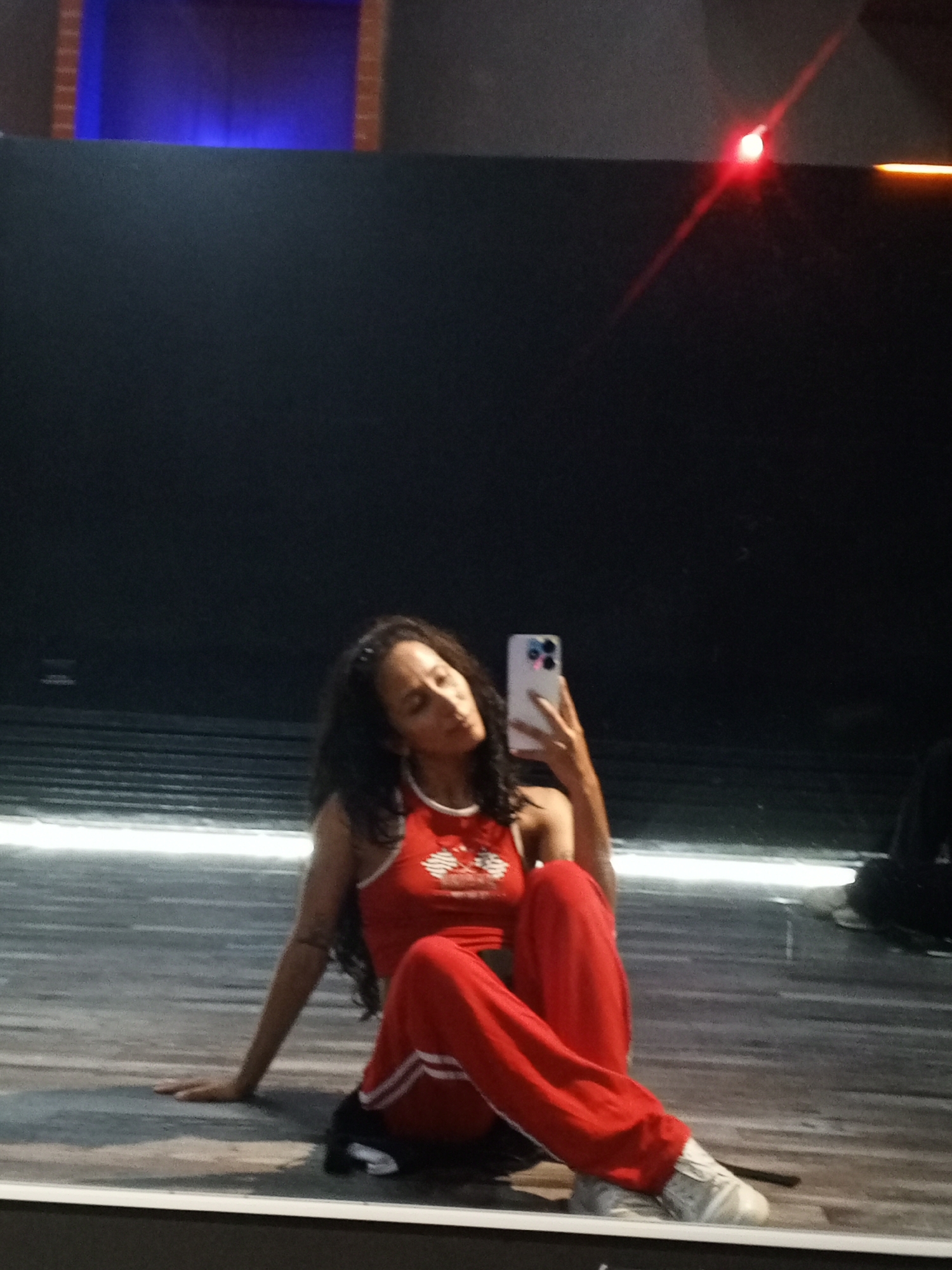 A woman is sitting on a reflective surface, holding a smartphone in her right hand, seemingly taking a selfie. She is wearing a red tank top with white detailing and matching red pants with white stripes down the sides. She also has on white sneakers. Her dark hair is down. The background features a dark wall with what appears to be a lit window in the upper left corner and a light reflection in the upper right corner. The reflective surface suggests she is in a mirrored room or in front of a large mirror. Due to the lack of any identifying characteristics, it is not possible to confirm this scene is in Pampatar, Venezuela.