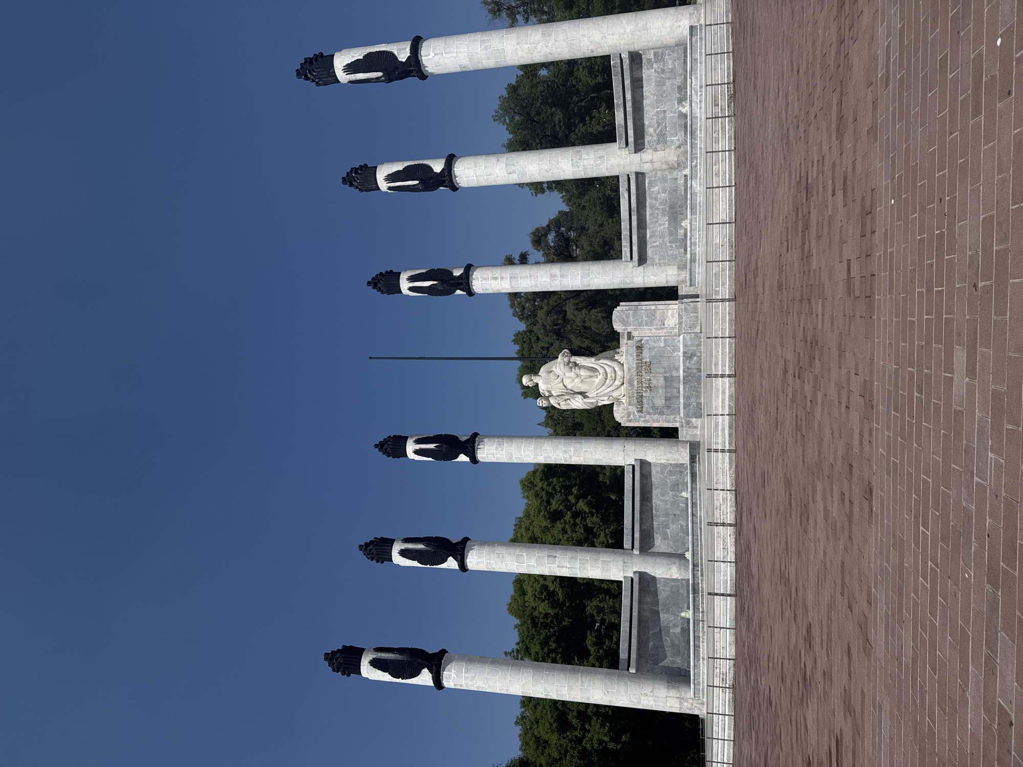 The image shows the Monument to the Heroes of the Restoration in Mexico City, Mexico. The monument features six white marble columns, each topped with a black eagle. At the back of the columns, there is a white marble statue. The monument is situated on a red brick ground. The sky is blue and clear. In the background, trees can be seen.