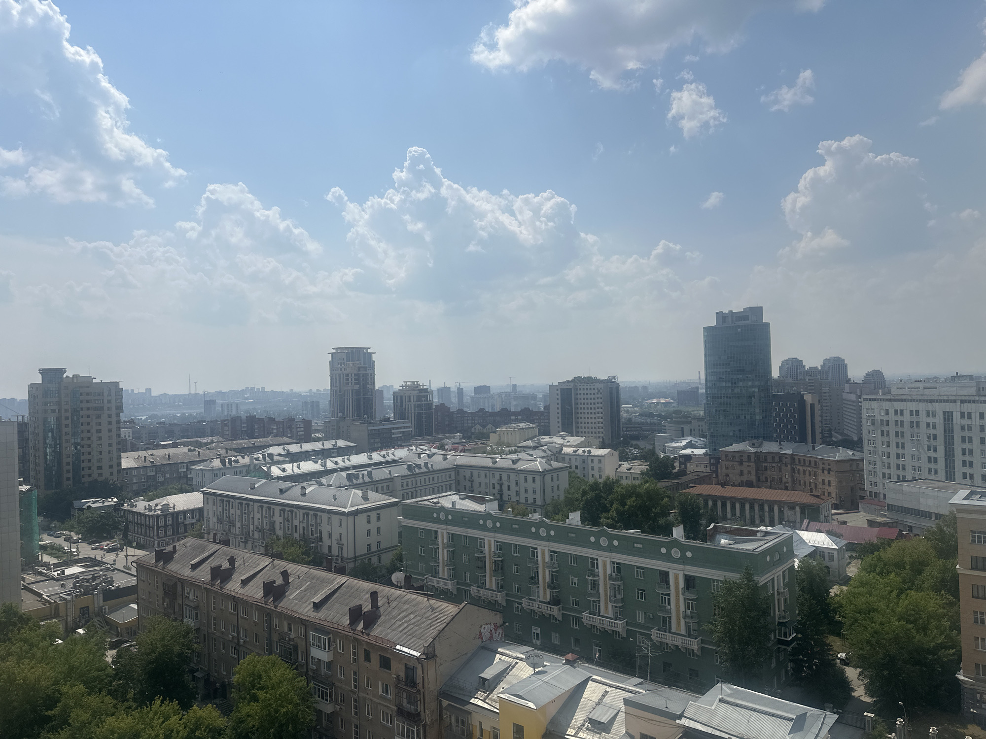 This is an aerial view of a dense urban environment in Novosibirsk, Russia, under a bright, slightly hazy sky with scattered cumulus clouds. The image captures a mix of architectural styles, predominantly residential buildings, ranging from older, lower-rise structures with pitched, dark roofs to more modern, taller apartment blocks with flat roofs.
In the foreground, a multi-story apartment building with a dark, sloped roof dominates the lower left portion of the frame. To its right, another apartment building features a distinctive green facade with white trim and a pattern of windows and balconies. Further back, rows of similar apartment buildings, many with light-colored facades, extend into the mid-ground. Some of these buildings exhibit classical architectural elements.
The background reveals a skyline punctuated by several modern, taller buildings, including glass-fronted skyscrapers and more traditional rectangular high-rises. The distant cityscape appears somewhat obscured by haze, suggesting a level of atmospheric pollution or the distance itself.
Vegetation, primarily mature trees with dense green foliage, is interspersed throughout the urban landscape, particularly in courtyards and along streets. A few cars are visible on the streets, and what appear to be some small utility structures are present on rooftops.
The scene depicts a typical urban day, with no prominent human activity or specific actions captured. The focus is on the built environment and the sky above. The composition suggests the vantage point is from a high-rise building overlooking the city.