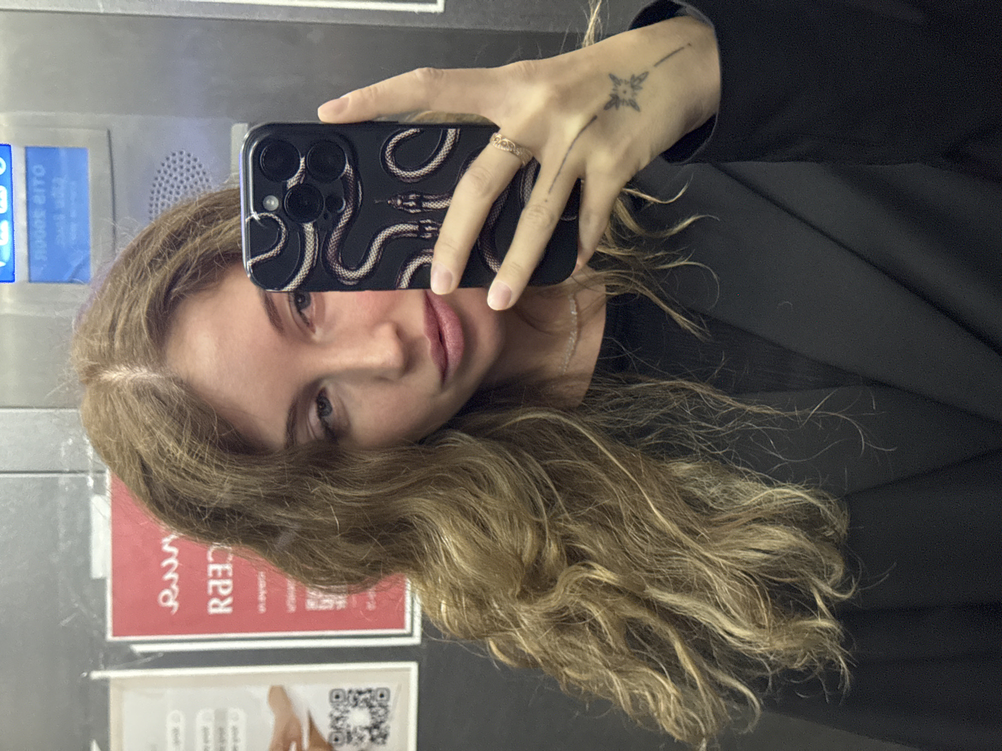A woman with long, wavy blonde hair is taking a selfie in an elevator. She is wearing a black blazer over a black top. Her right hand is holding a black smartphone with a snake design case. She is also wearing a ring on her right ring finger and has a tattoo on her right wrist. In the background, there are posters on the elevator wall. One poster is red with white text and appears to be in Russian. Another poster is white and has a QR code. The elevator control panel is visible at the top of the image, displaying "OTIS". The scene is indoors, in an elevator. The location is Chaikovsky, Russia.