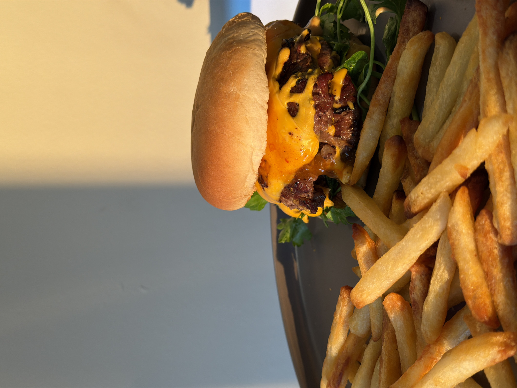 A close-up, eye-level shot shows a gourmet double cheeseburger and a pile of golden french fries on a dark grey plate, set against a light grey wall with a warm yellow glow from sunlight casting shadows. The burger, positioned on the right side of the frame, features a toasted bun, melted bright orange cheese cascading over two thick, grilled beef patties, and a visible layer of green arugula. The fries, filling the left side and bottom of the frame, are crisp and appear seasoned, with some showing darker, fried edges. The lighting suggests late afternoon or early morning, possibly near a window. The overall scene evokes a feeling of a casual, high-quality meal, potentially in a restaurant or home setting. The location is not specified in the image.