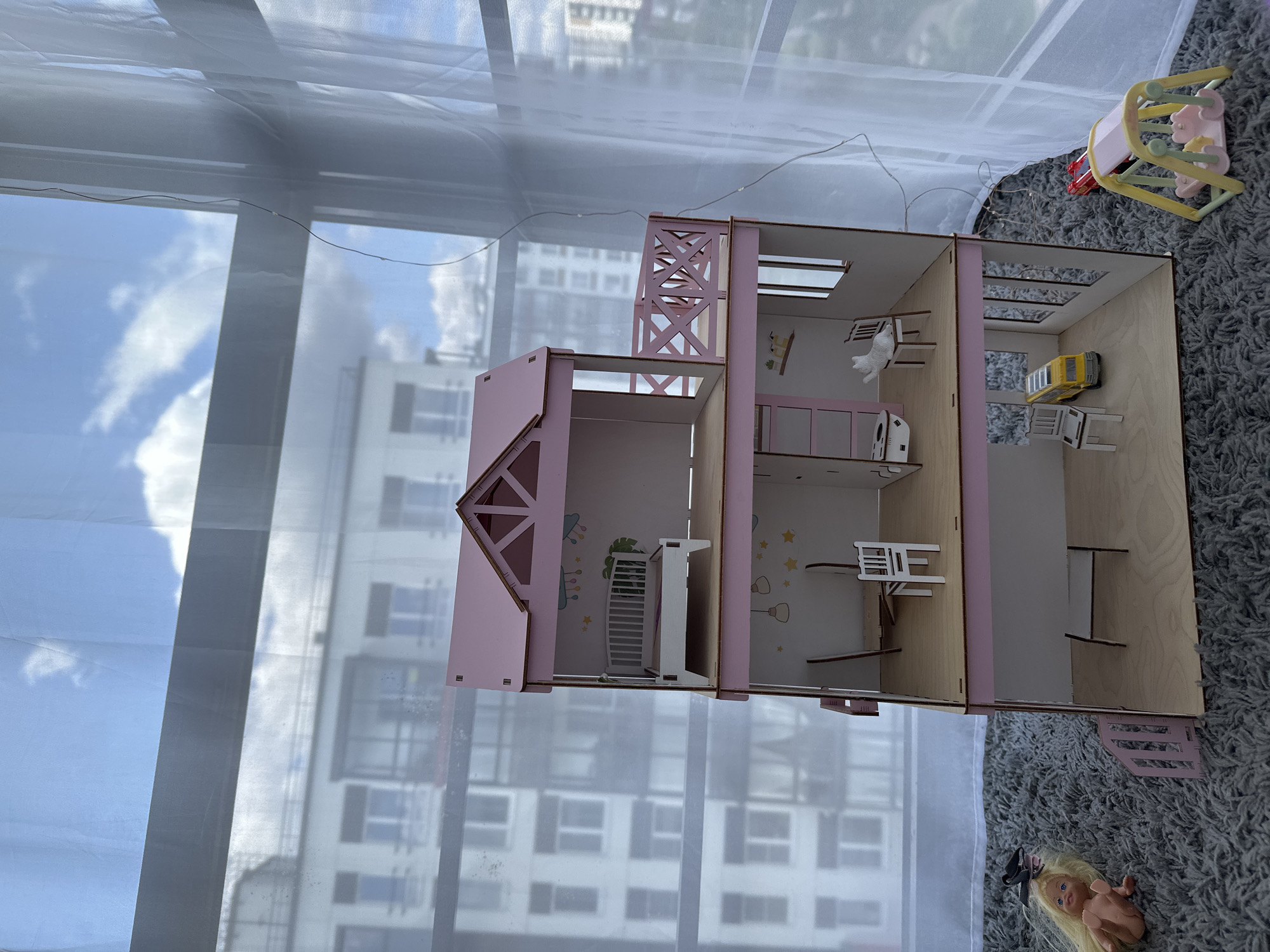 A three-story pink and wood dollhouse is situated on a grey carpet in Vsevolozhsk, Russia, in front of a window with sheer white curtains and a view of apartment buildings and a cloudy sky. The dollhouse is furnished with a bed, chairs, a toilet, and a small yellow car. To the left of the dollhouse, a doll with blonde hair lies on the carpet. To the right, a miniature swing set with two pink figures is visible. A red toy car is positioned on the carpet near the swing set. A string of small lights is draped along the curtain. The lighting in the image is natural and diffused through the window.