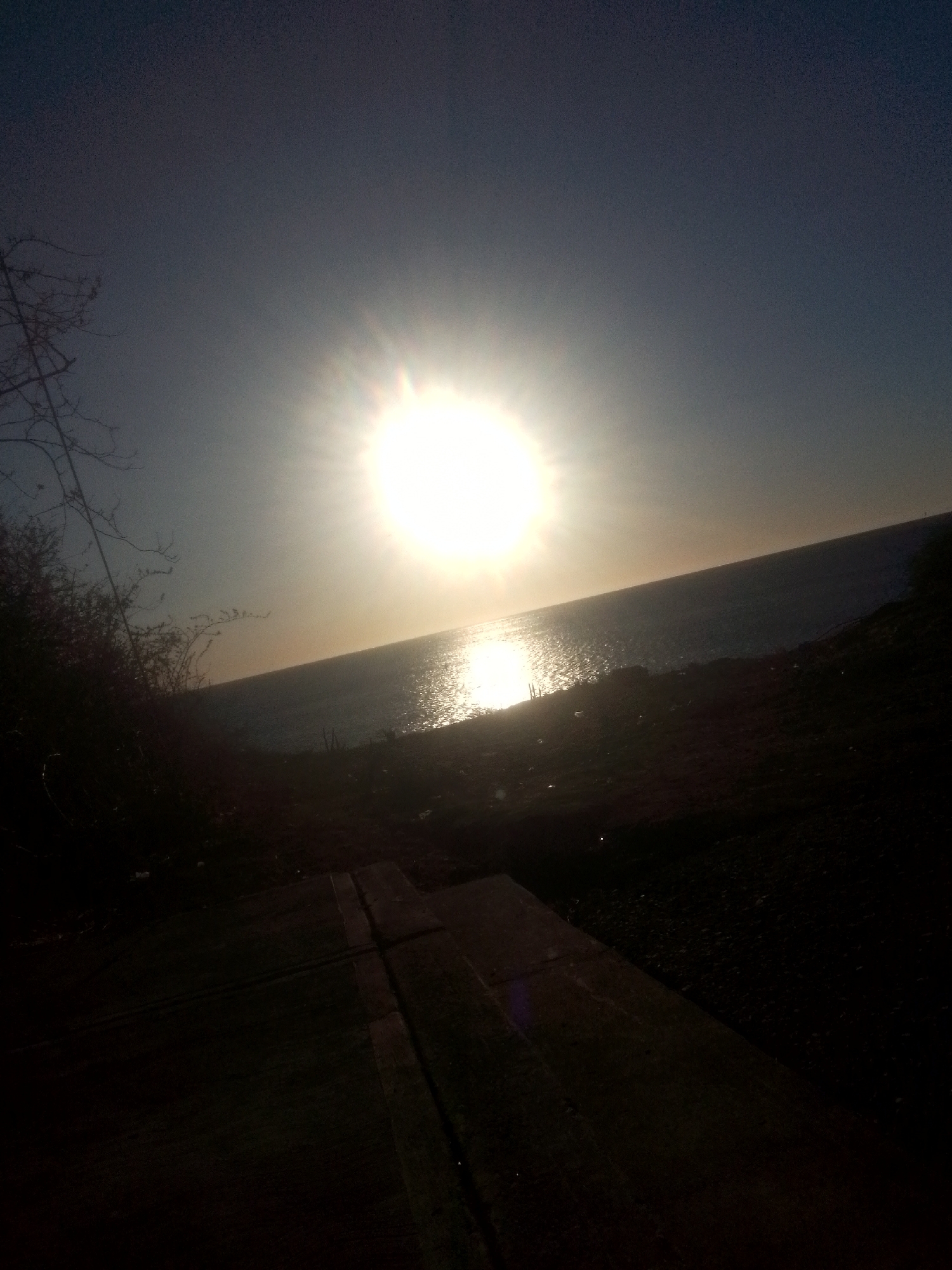 The image depicts a sunset over the ocean, possibly in Juan Griego, Venezuela. The sun is a bright orb in the center of the frame, with its reflection visible on the water's surface. The horizon is a straight line separating the sky and the sea. The foreground is dark and appears to be a pathway or deck, possibly leading towards the water. A cluster of vegetation is visible to the left of the path.