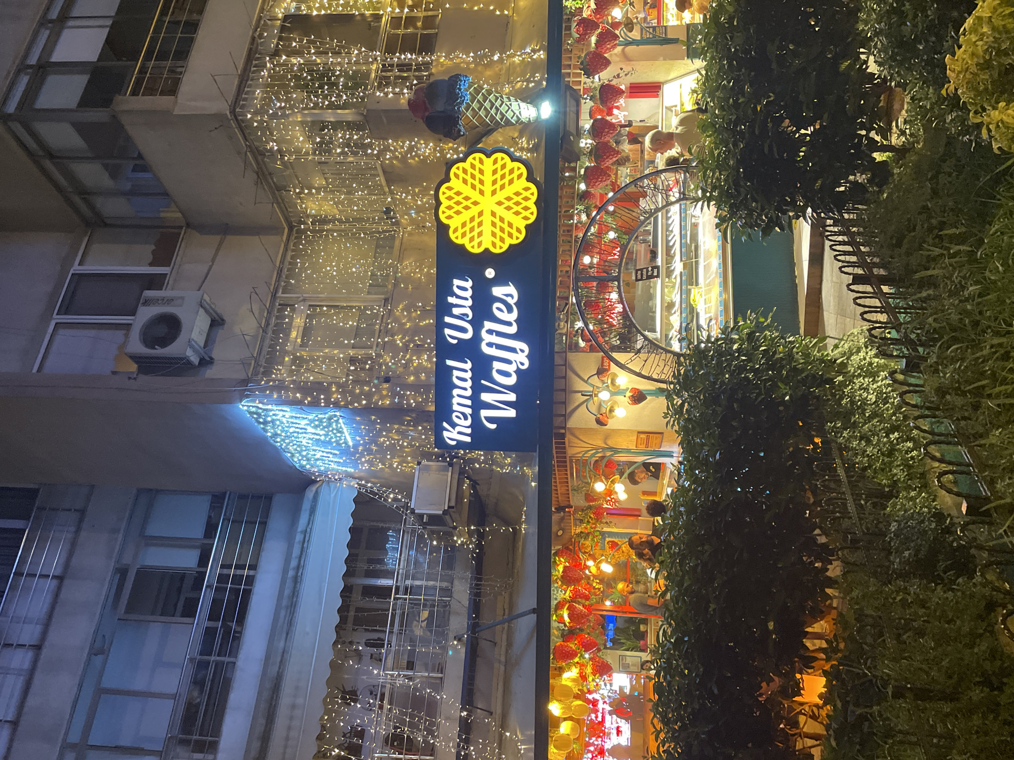 This image captures the exterior of a waffle shop called "Kemal Usta Waffles" at night in Kadıköy, Turkey. The shop is adorned with numerous twinkling lights, creating a festive ambiance. A prominent sign above the entrance displays the shop's name in illuminated lettering. To the right of the sign, a large, stylized waffle graphic is visible, and next to that, a decorative ice cream cone complete with scoops of ice cream.
Below the sign, the shop's interior is visible, showcasing display cases filled with various pastries and desserts. Several customers can be seen inside the shop, some interacting with staff behind the counter, while others appear to be browsing the offerings.
Outside the shop, a line of lush green bushes partially obscures the view of the sidewalk and entrance. A decorative metal fence runs along the front of the bushes. The overall scene is brightly lit by a combination of the shop's signage, interior lights, and decorative string lights, all set against the backdrop of the urban night. The building itself appears to be a multi-story structure, with balconies and windows visible on the upper levels.