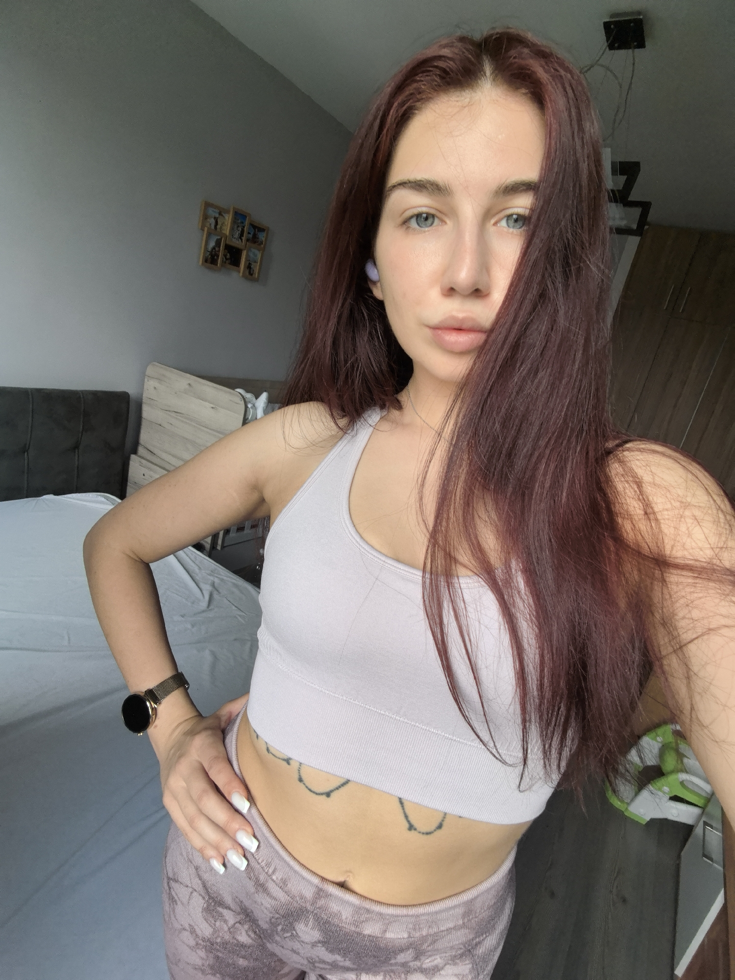 A young woman with long, dark reddish-brown hair and blue eyes poses for a selfie indoors, likely in a bedroom in Stara Zagora, Bulgaria, during the daytime. She stands with her left hand on her hip, looking directly at the camera with a neutral, confident expression. She wears a light purple ribbed sports bra and patterned purple athletic leggings. A small, chain-like tattoo is visible on her lower abdomen. She has a purple wireless earbud in her left ear and a gold-colored smartwatch with a mesh band on her left wrist. Her fingernails are painted white.
The background shows a simple, well-lit room. To her left, a dark grey upholstered headboard and white bedsheets are visible. Behind her, a light grey wall features a cluster of small framed pictures. To her right, part of a wooden bed frame, dark wooden wardrobe doors, and a modern black ceiling light fixture are seen. On the dark wooden floor in the lower right corner, a pair of green and white athletic shoes rests. The natural lighting suggests it is daytime. No discernible text is visible in the image.