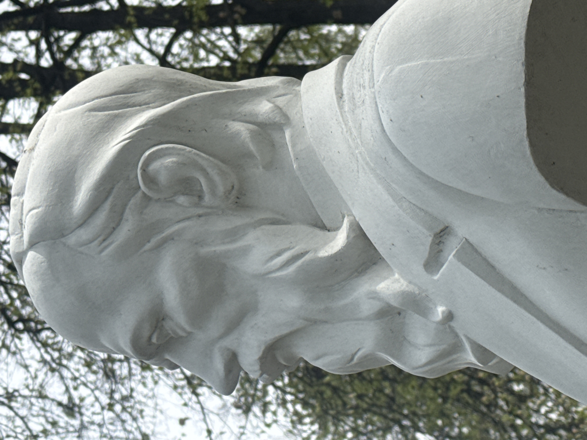 Close-up of a white marble statue's head and part of the upper body. The statue depicts a man with a shaved head and a visible ear, clad in what appears to be a formal shirt with a high collar. His head is bowed. The background shows blurry tree branches and sky, typical of an outdoor environment. This statue is part of the monument to Vasily Tatishchev and Georg Wilhelm de Gennin, located in Yekaterinburg, Russia.