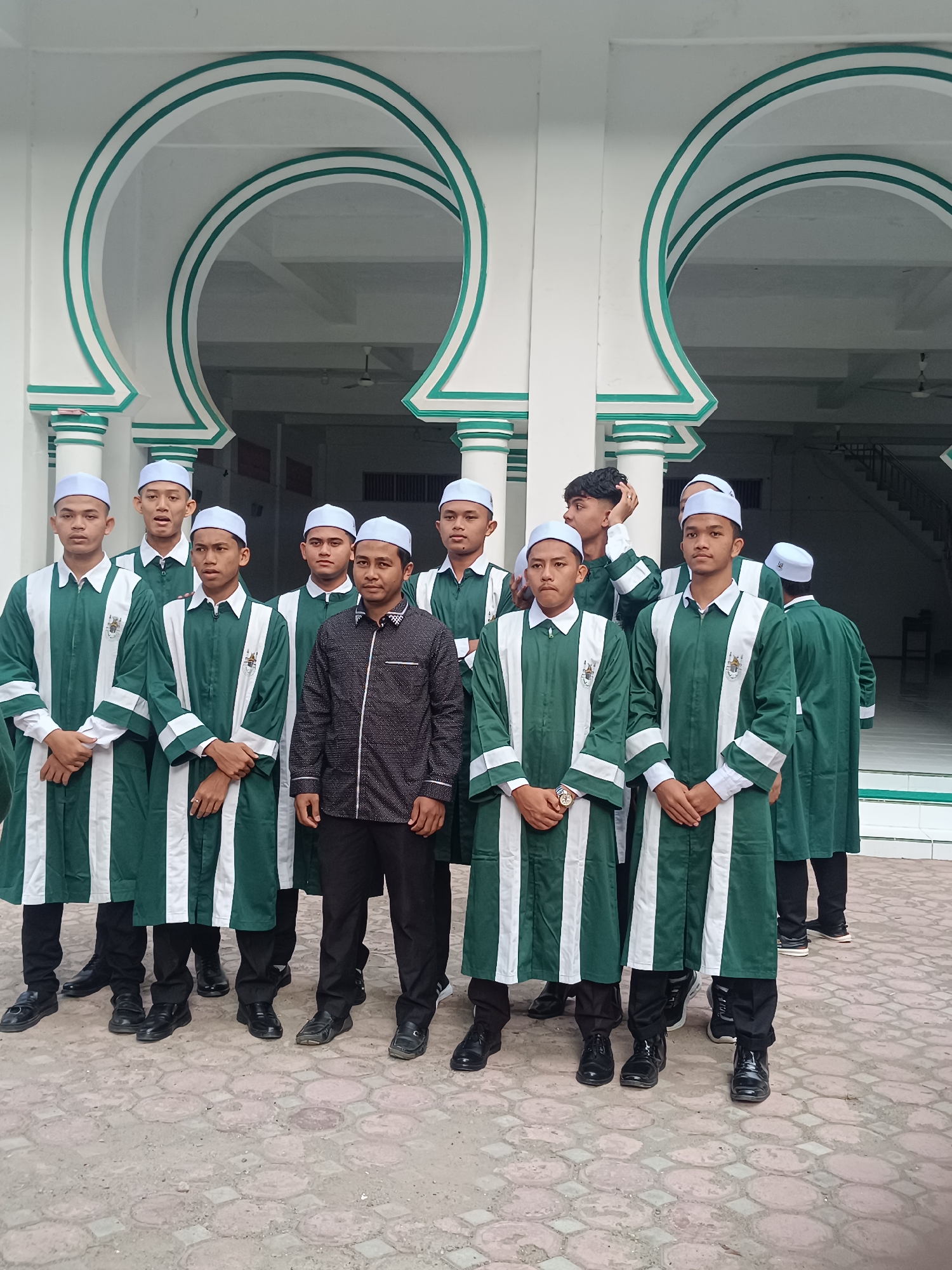 A group of 11 young men are standing in front of a white building with green trim and arched entrances. The men are wearing matching green and white robes and white skullcaps, except for one man in the center of the group who wears a black patterned shirt and black pants. All the men are wearing black shoes. The location is likely in Lhokseumawe, Indonesia, based on the visual style of the building and clothing. The men are posing for a photograph, with some looking directly at the camera and others looking elsewhere.