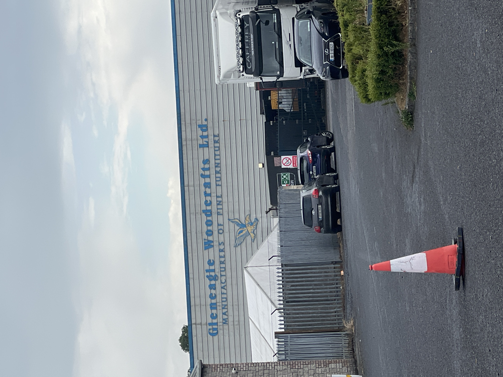 The image shows the exterior of a large industrial building with "Gleneagle Woodcrafts Ltd. Manufacturers of Fine Furniture" written in blue lettering across the facade. Below the lettering, a stylized depiction of a bird in flight is visible. In front of the building, there is a white tent structure and a metal fence. Several vehicles are parked in the foreground, including a dark-colored Lexus, a blue hatchback, and a grey hatchback. A large white truck with "CAFFREY" written on it is also present. A red and white traffic cone is in the lower left corner of the image. The sky is overcast.