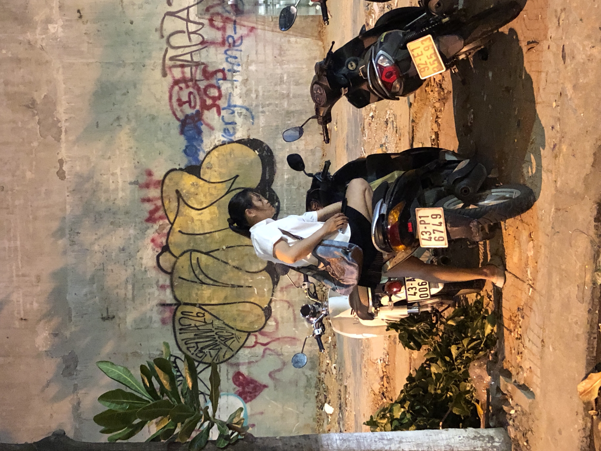In an outdoor setting at night, a young woman wearing a white collared shirt and dark skirt is perched on the seat of a black scooter, leaning to her right with her left leg extended. She has a brown shoulder bag. Two other scooters are parked nearby: a white scooter to her left and another black scooter to her right. The scooters are parked on a paved surface next to a rough concrete wall adorned with graffiti. To the left of the scooters and woman, a tree with green foliage is visible, and a pile of leafy branches lies on the ground. The overall scene suggests a street or alleyway in a city. The license plates visible on the scooters read "43-A 006" and "43-P1 6749" on the left and middle scooters respectively, and "92-E1 355.93" on the right scooter. Based on the license plate prefixes "43" which indicates Đà Nẵng province, it is highly probable that this scene is located in the city of Đà Nẵng, Vietnam.