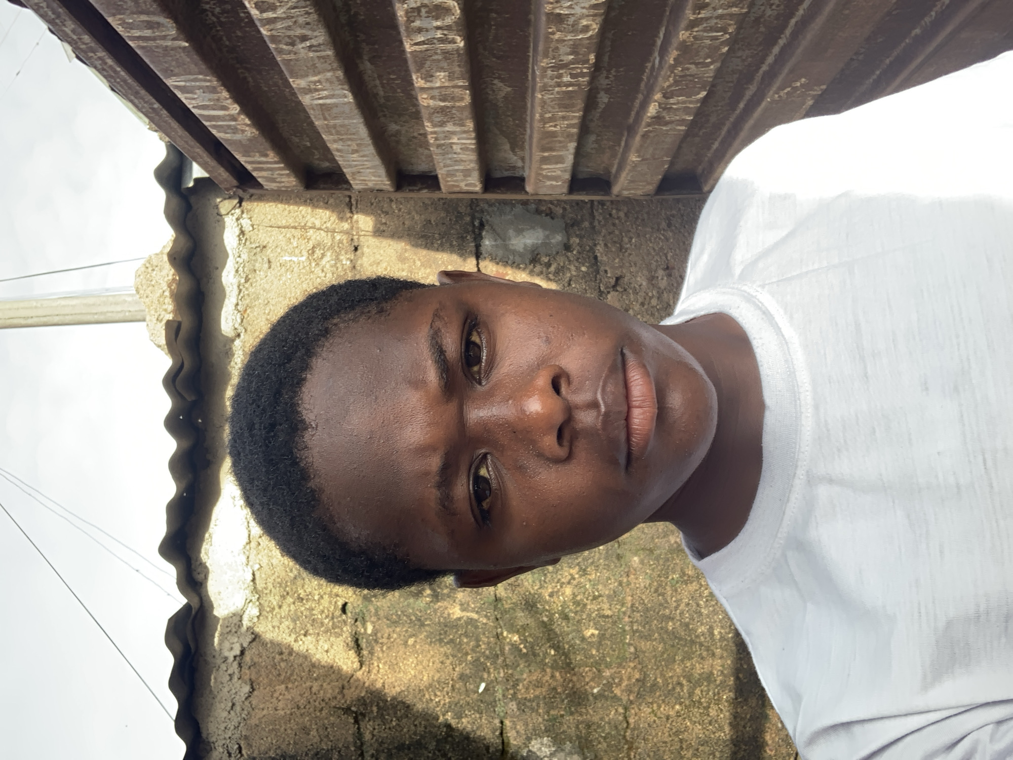 A young person with dark skin and short, dark hair poses for a close-up, eye-level selfie outdoors. The person wears a white t-shirt. Behind them, a textured, light-colored wall with shadows from overhead corrugated material stretches across the background. To the right, a rusty metal gate with horizontal slats is visible. The overall environment suggests an outdoor urban setting, possibly in Jos, Nigeria, indicated by the specific details and cultural context.
