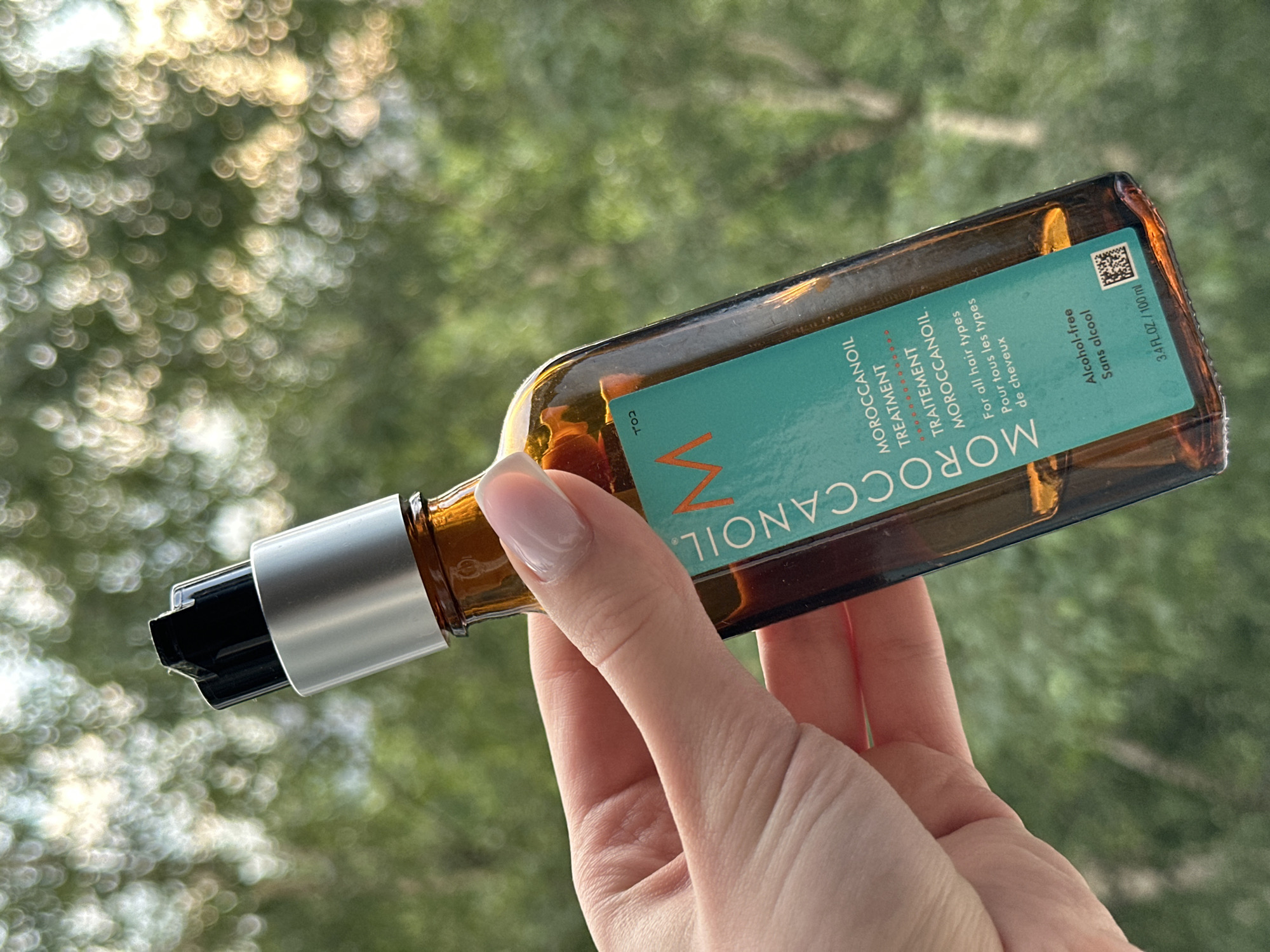 A hand with light skin and a sheer, pale pink manicure is holding a small, amber-colored glass bottle of Moroccanoil Treatment against a softly blurred background of green foliage and bright, bokeh-like circles of light, possibly indicating an outdoor setting with dappled sunlight. The bottle has a silver pump dispenser with a black plastic cap. A turquoise label is visible on the bottle, featuring the Moroccanoil logo and text in English and French, including "MOROCCANOIL TREATMENT," "TRAITEMENT MOROCCANOIL," "For all hair types / Pour tous les types de cheveux," "Alcohol-free / Sans alcool," and "3.4 FL.OZ / 100 ML." A QR code is also present on the label. The scene does not contain any identifiable landmarks or indicators of its specific location within Moscow, Russia.