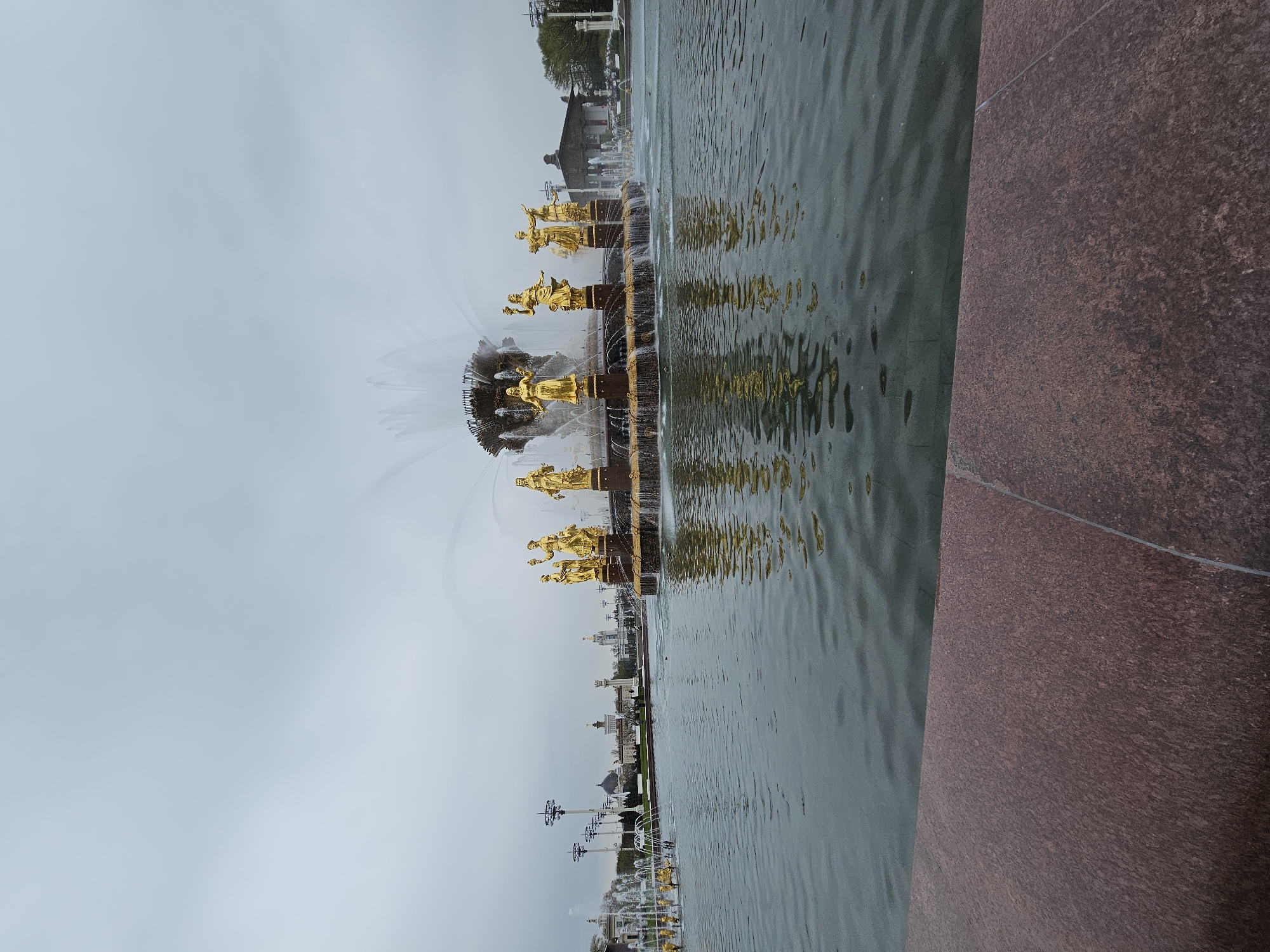 The image depicts the Friendship of Nations Fountain in Moscow, Russia. The fountain is adorned with gilded statues of women representing the different republics of the Soviet Union. Water is flowing from the fountain into a pool. The sky is overcast and gray. A red granite structure is visible on the right. Buildings can be seen in the distance.