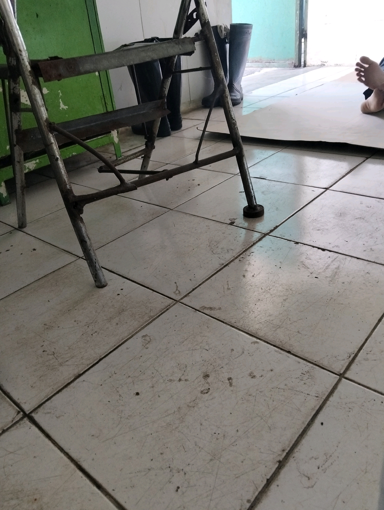 The image depicts an interior scene on a tiled floor, likely in Tanggulangin, Indonesia. In the foreground, a rusted metal folding stool with wooden steps is positioned. Behind the stool, a pair of dark rubber boots stands upright. To the right, a person's bare foot is visible, with toes extended. The environment appears to be an indoor space with a green cabinet or wall on the left and a light-colored floor. Sunlight streams in from the right, creating shadows on the floor. The overall impression is that of a utilitarian or storage area.
