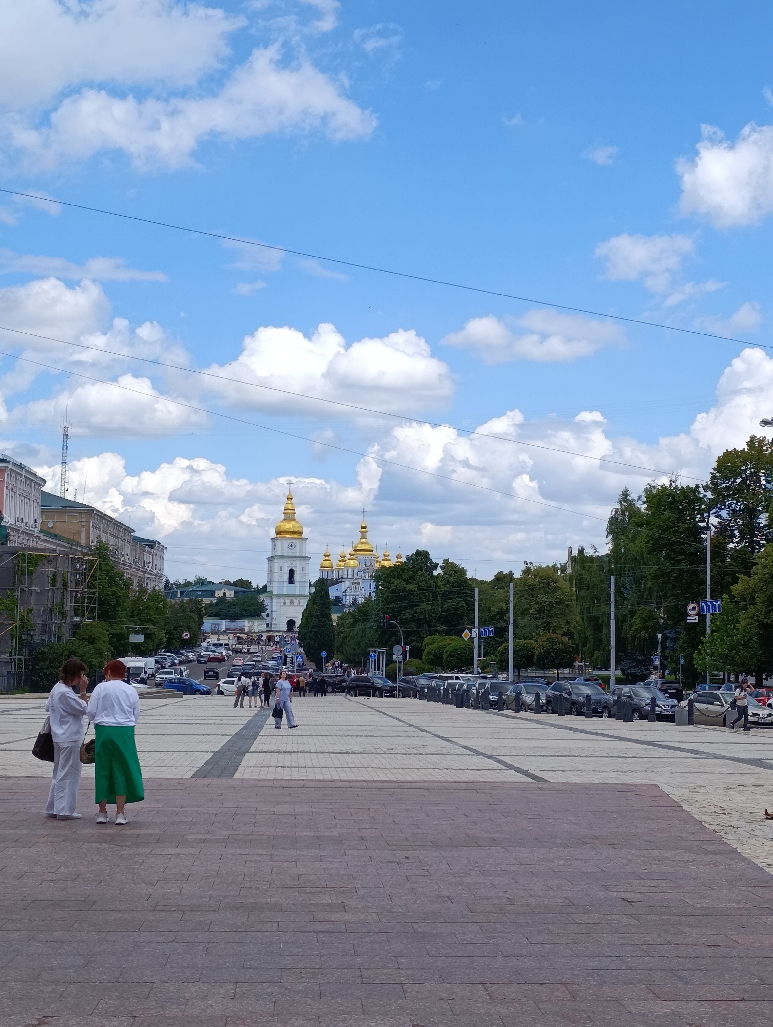 The image depicts a wide paved plaza in Kyiv, Ukraine, likely St. Sophia's Square, looking towards St. Michael's Golden-Domed Monastery. In the foreground, two women are standing on a set of steps, one in white attire and the other in white and a green skirt. Beyond them, the plaza opens up to a street lined with parked cars and trees. Further in the distance, the iconic golden domes of St. Michael's Monastery are prominently visible against a bright blue sky dotted with fluffy white clouds. The architecture of surrounding buildings, including a building with scaffolding, suggests an urban setting within Kyiv. The scene appears to be from a daytime event, with people walking and cars moving through the street.